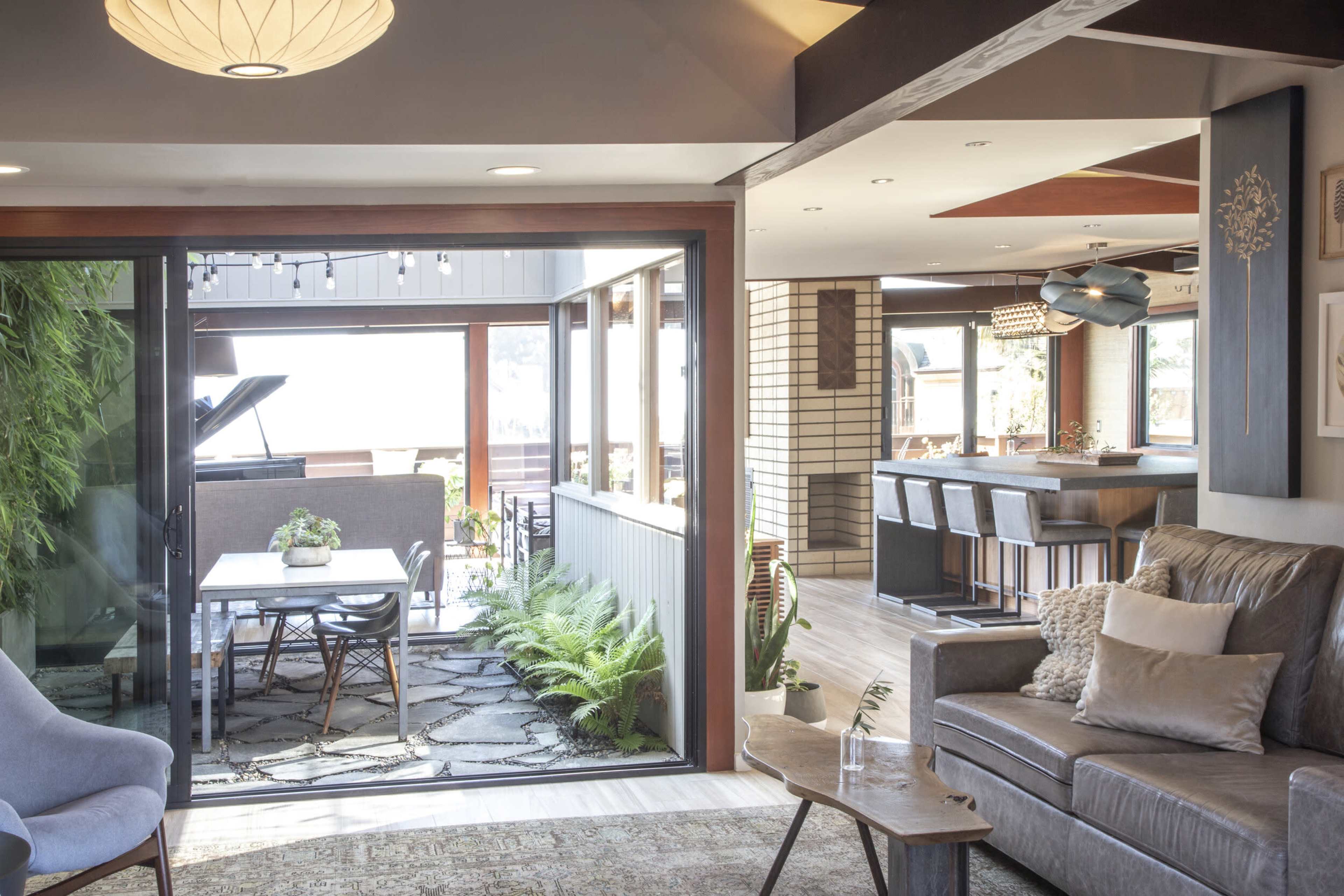 The image shows an open-concept living space that transitions into an outdoor seating area, featuring a modern design with a mix of indoor plants, furniture, and a dining table.