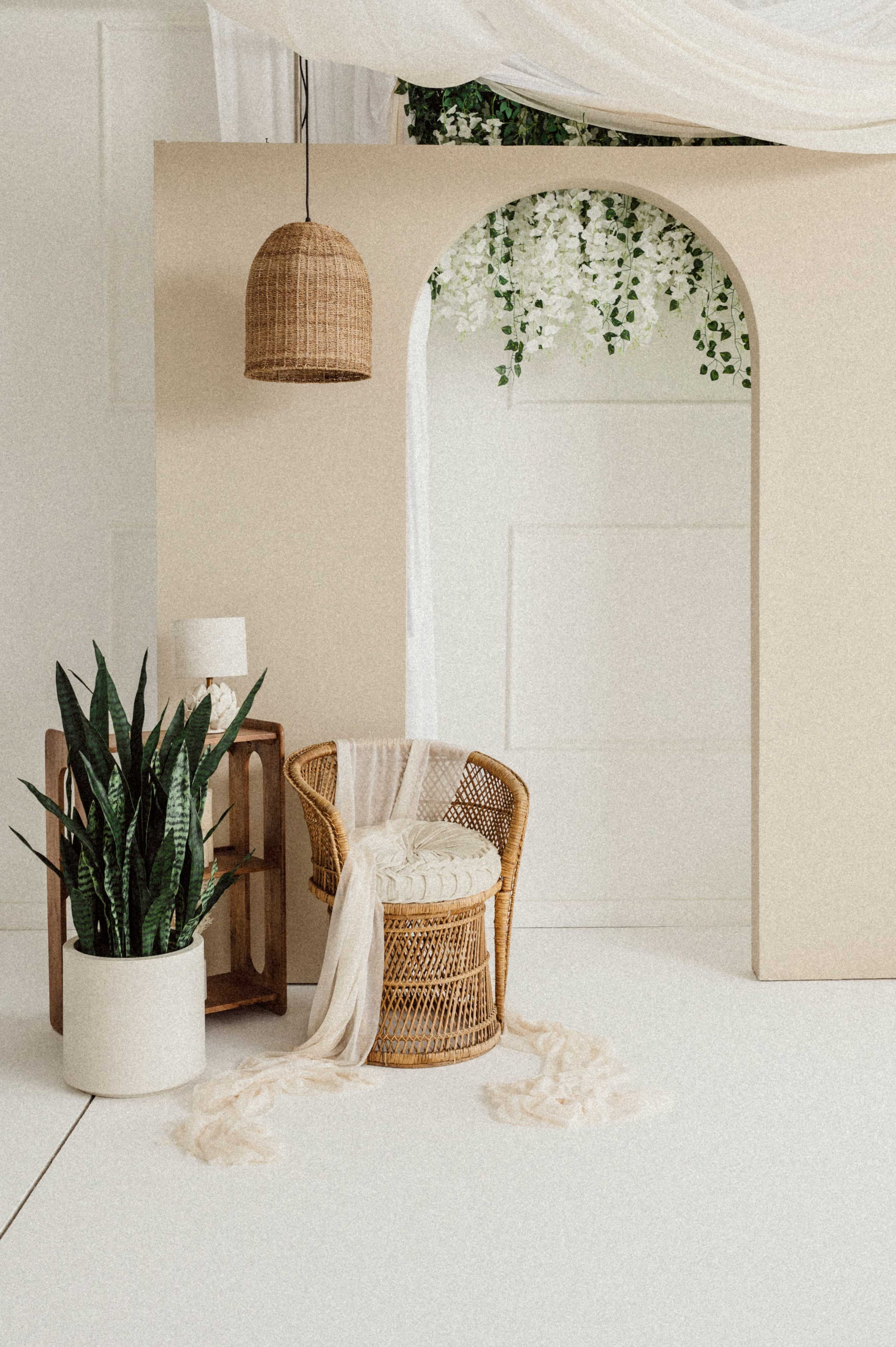 The image features a minimalistic interior scene with a woven chair, a plant in a white pot, and a decorative arch adorned with greenery, all set against a light-colored backdrop.