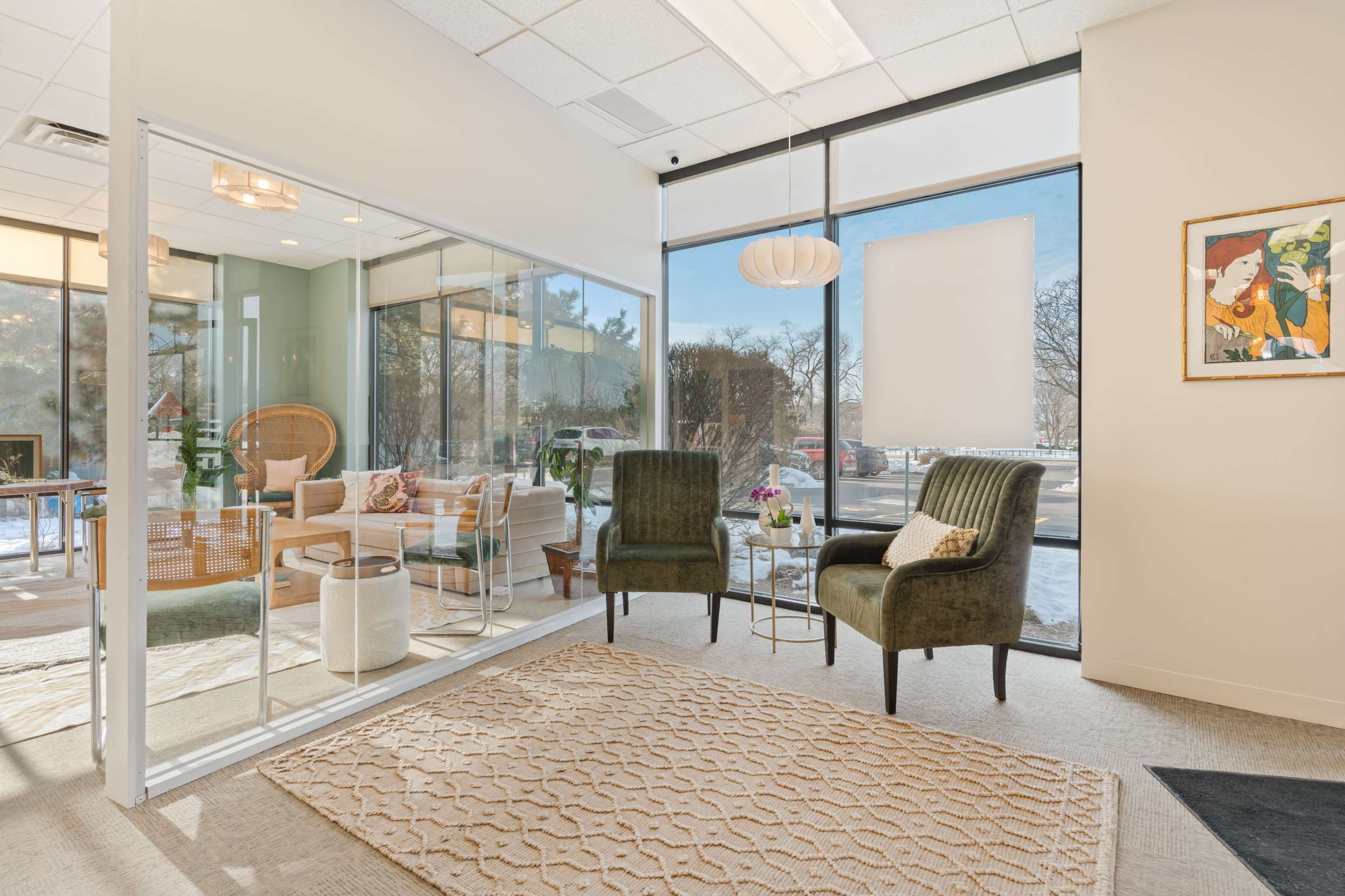 The image shows a modern office waiting area with two green chairs, a small glass table, and large windows providing natural light.