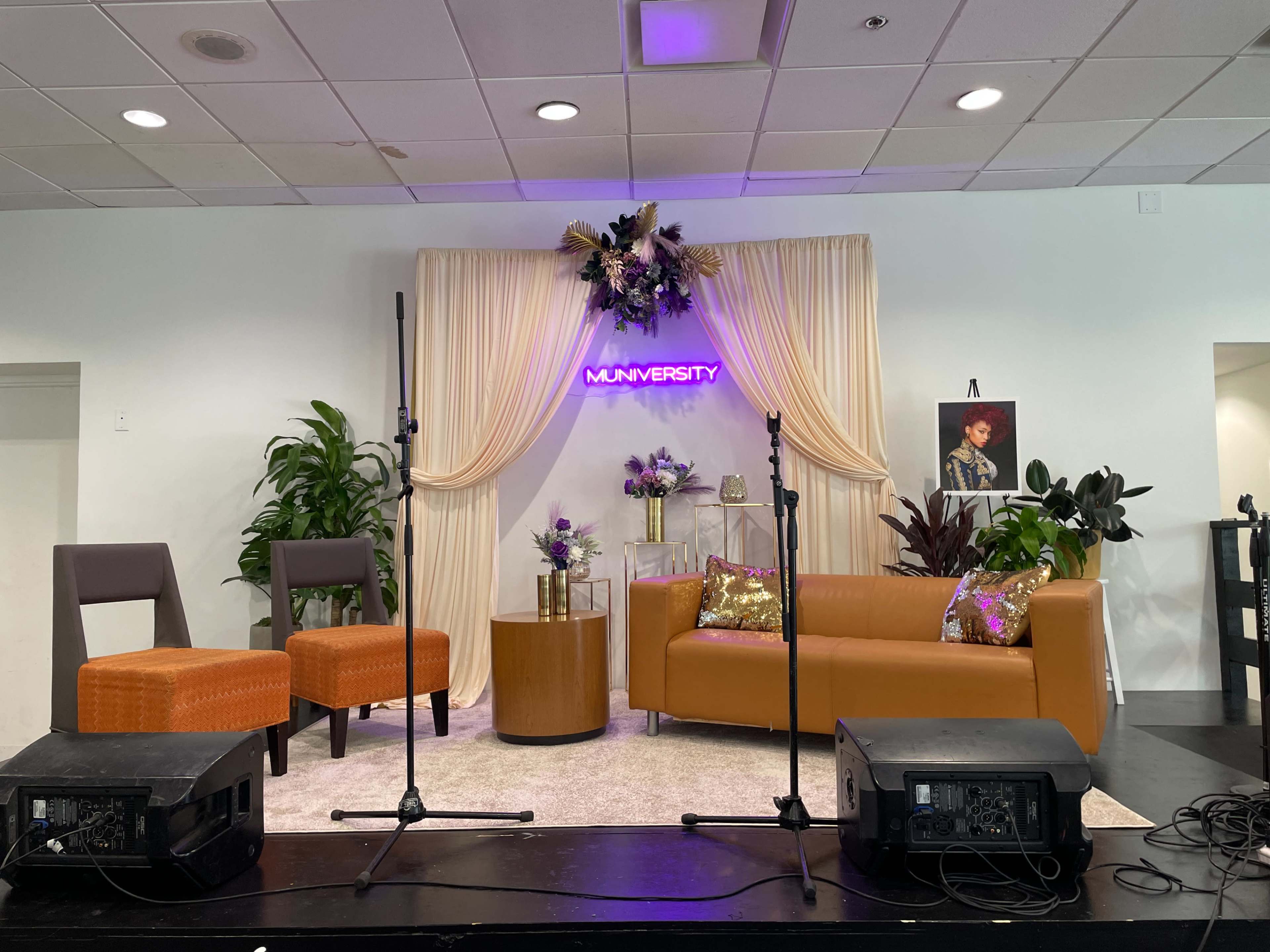 The image shows a stage setup with two chairs, a stylish sofa, a decorative backdrop featuring plants and a sign that reads "MUNVERSITY," alongside microphones and speakers.