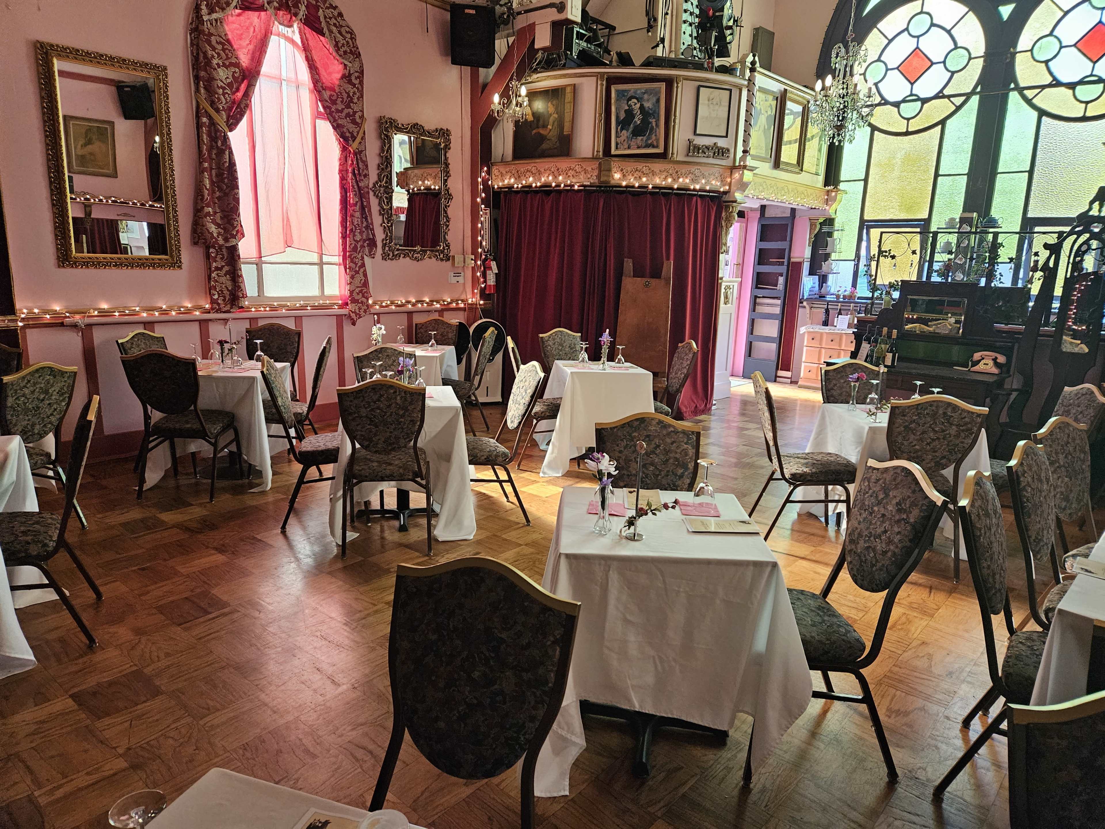 A dining area with several neatly set tables and chairs, surrounded by ornate mirrors and draped curtains, within a venue featuring large stained-glass windows.