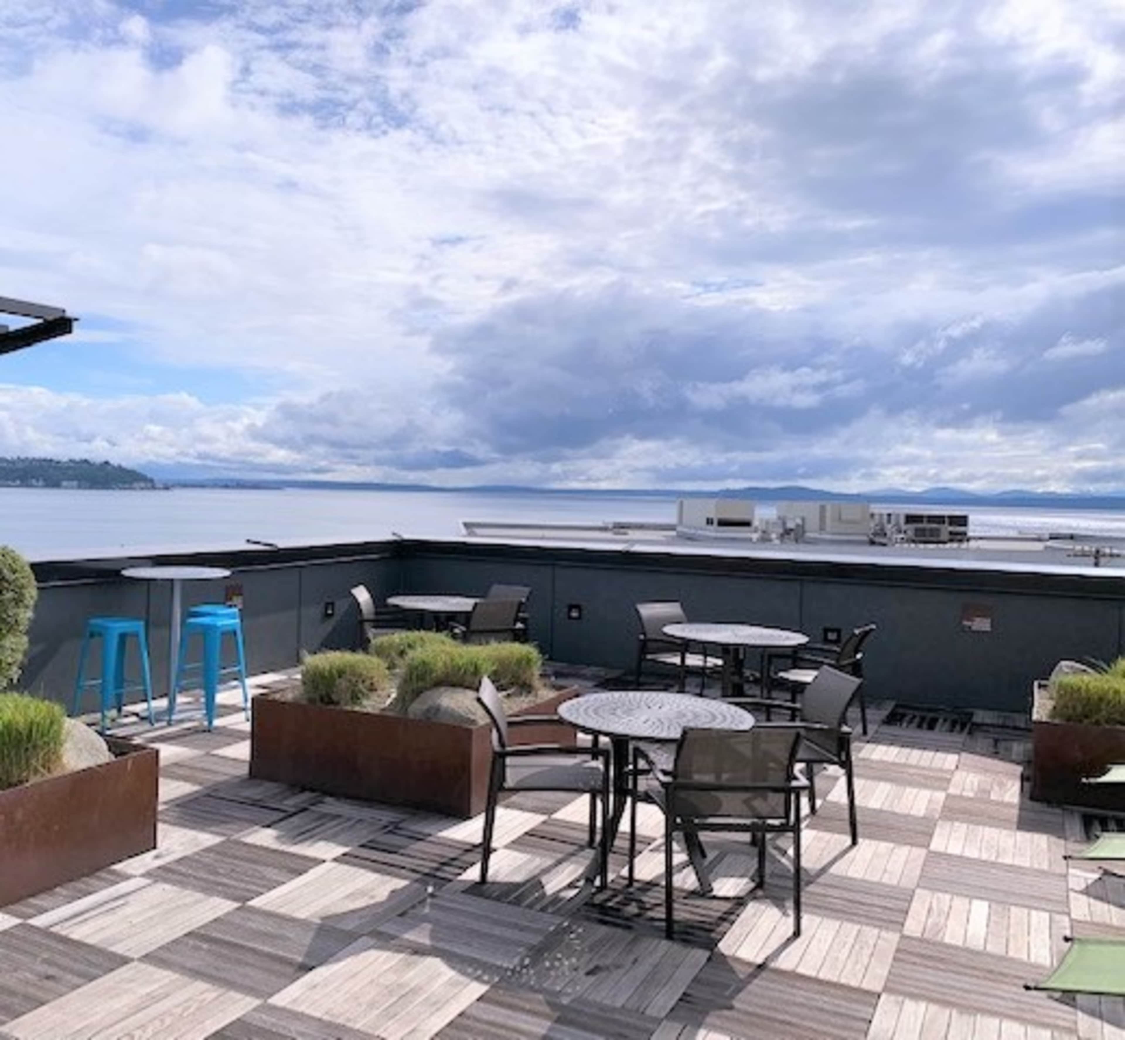 A rooftop terrace features several tables and chairs overlooking a body of water under a partly cloudy sky.