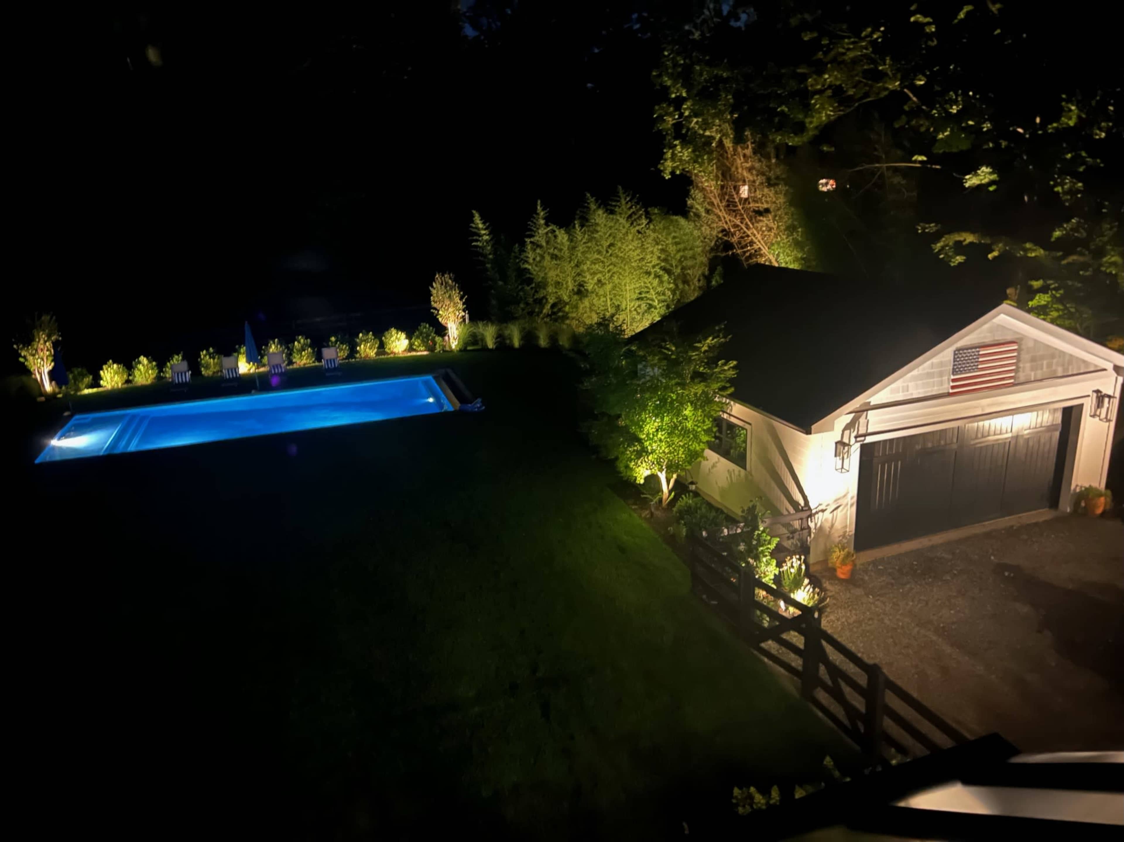 A well-lit backyard at night featuring a swimming pool and a detached garage with an American flag.