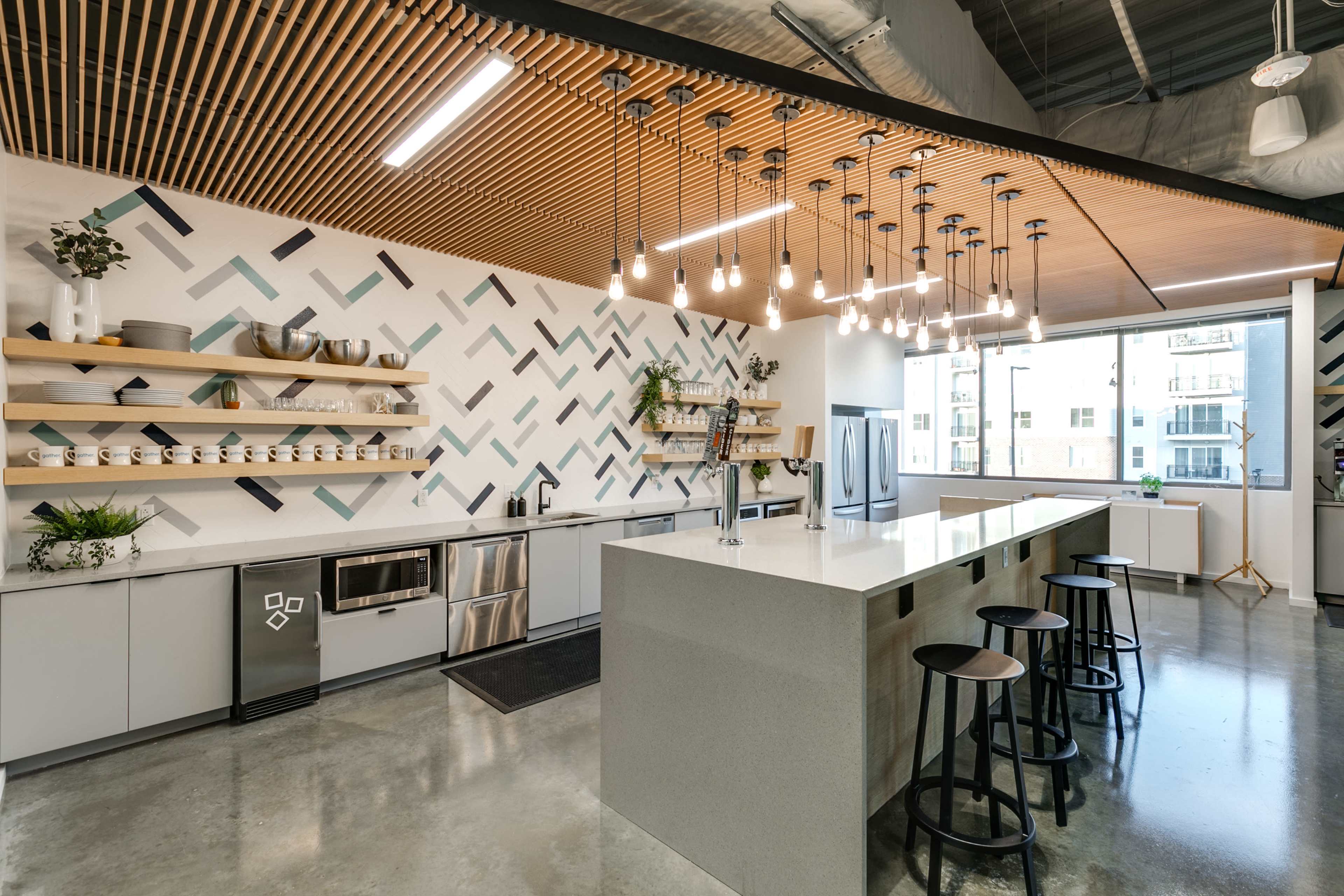 The image shows a modern kitchen featuring a large island with seating, open shelving on the walls, and hanging light fixtures, set against a decorative patterned backsplash.