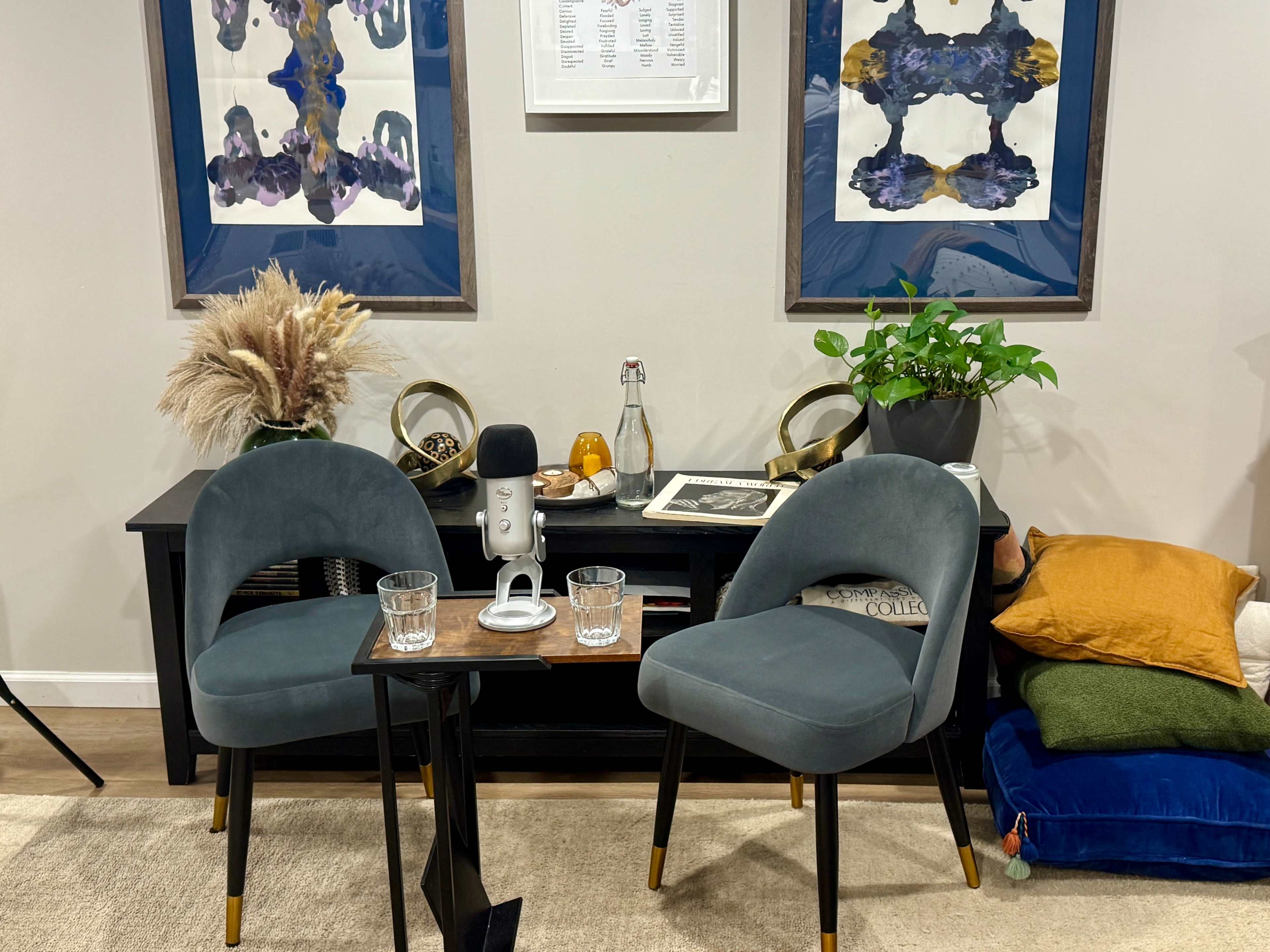 The image shows a cozy interior space with two gray chairs placed around a small table, which holds a microphone and two glasses, alongside a decorative display of framed artwork and various plants.