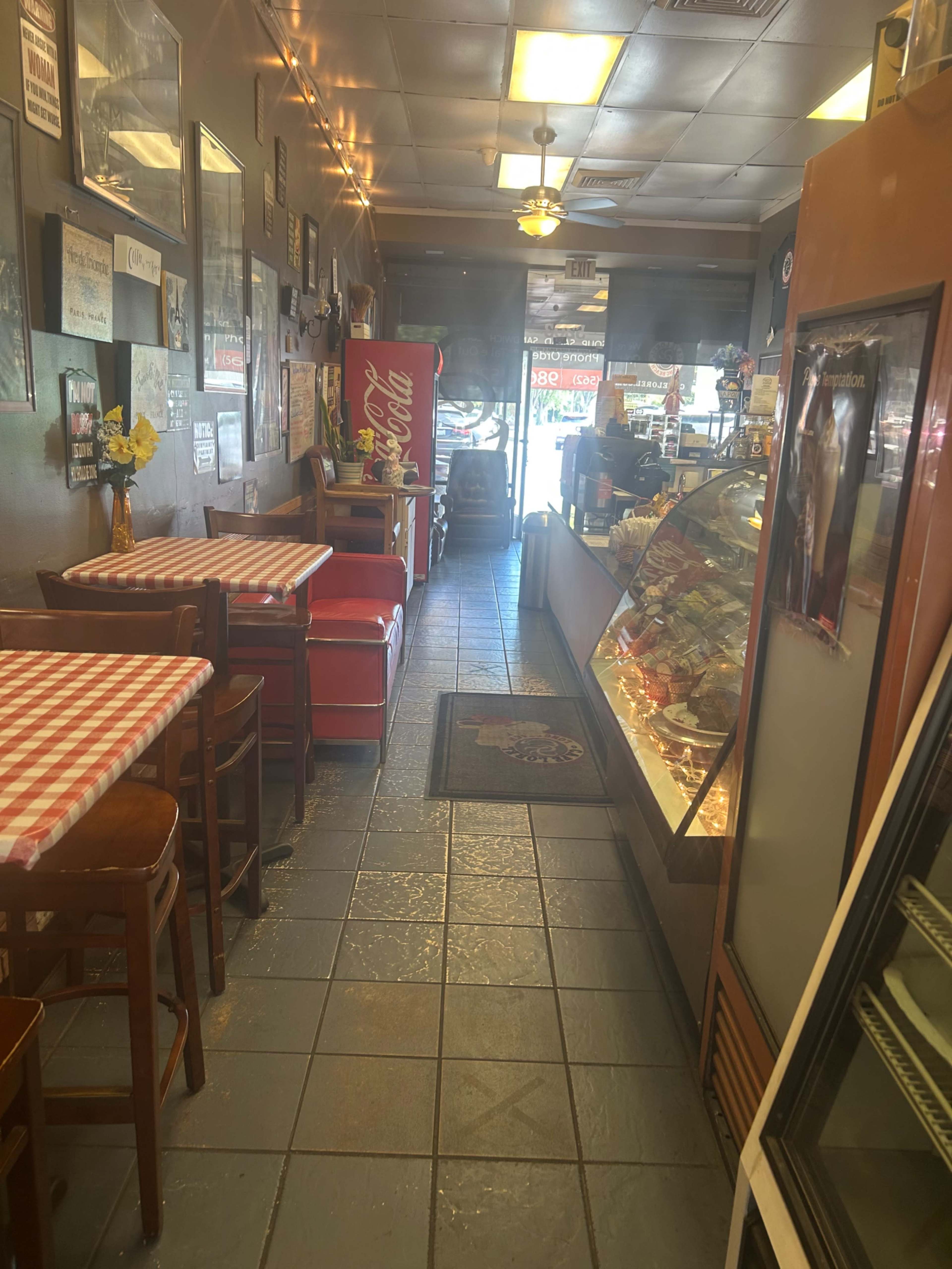 The image shows a café interior featuring two small tables with checkered tablecloths, a counter displaying pastries, and a red Coca-Cola vending machine along a narrow walkway.
