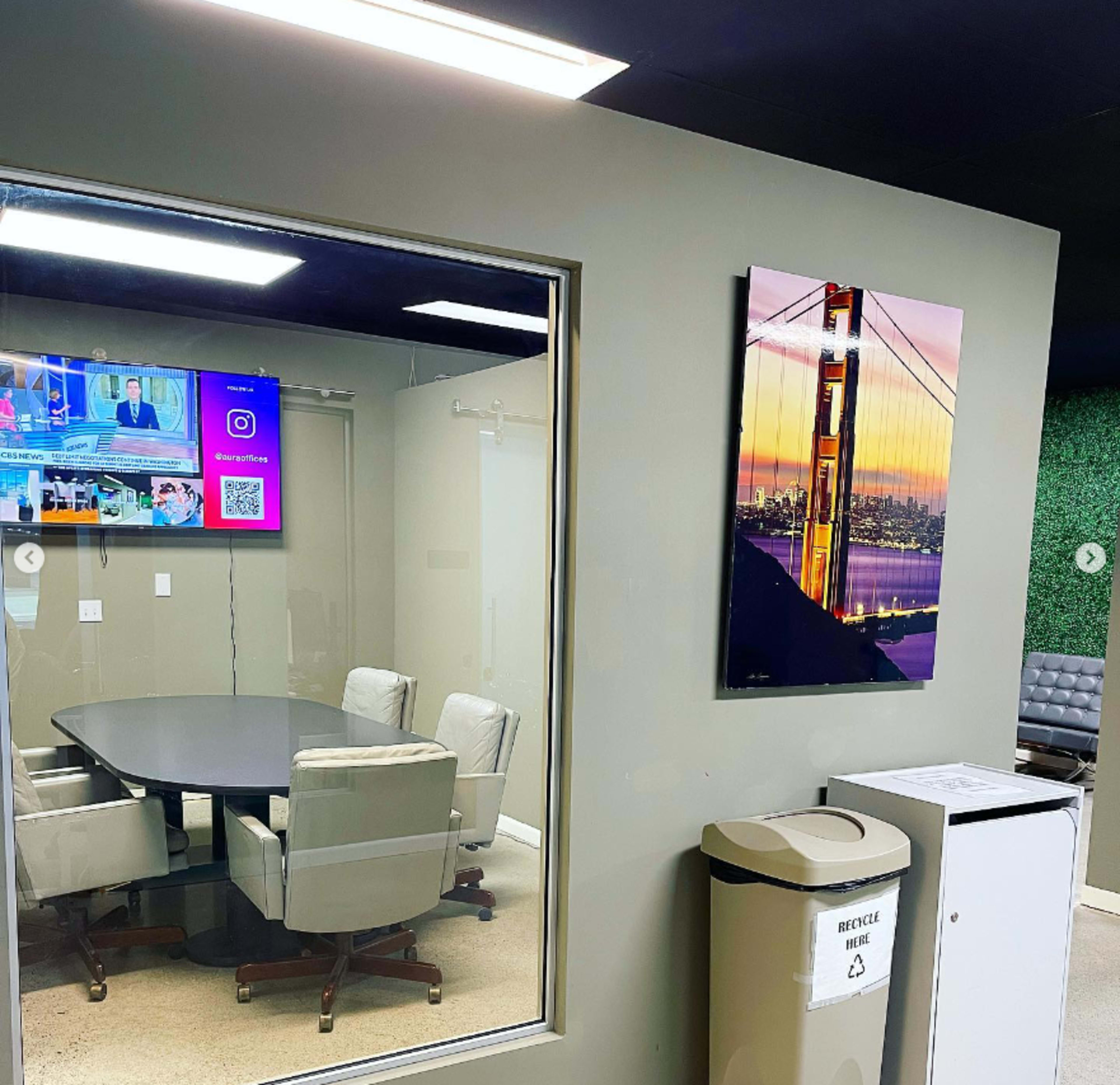 A conference room with a round table and ergonomic chairs is visible through a glass wall, while a TV shows news and a colorful image of the Golden Gate Bridge hangs on the adjacent wall.