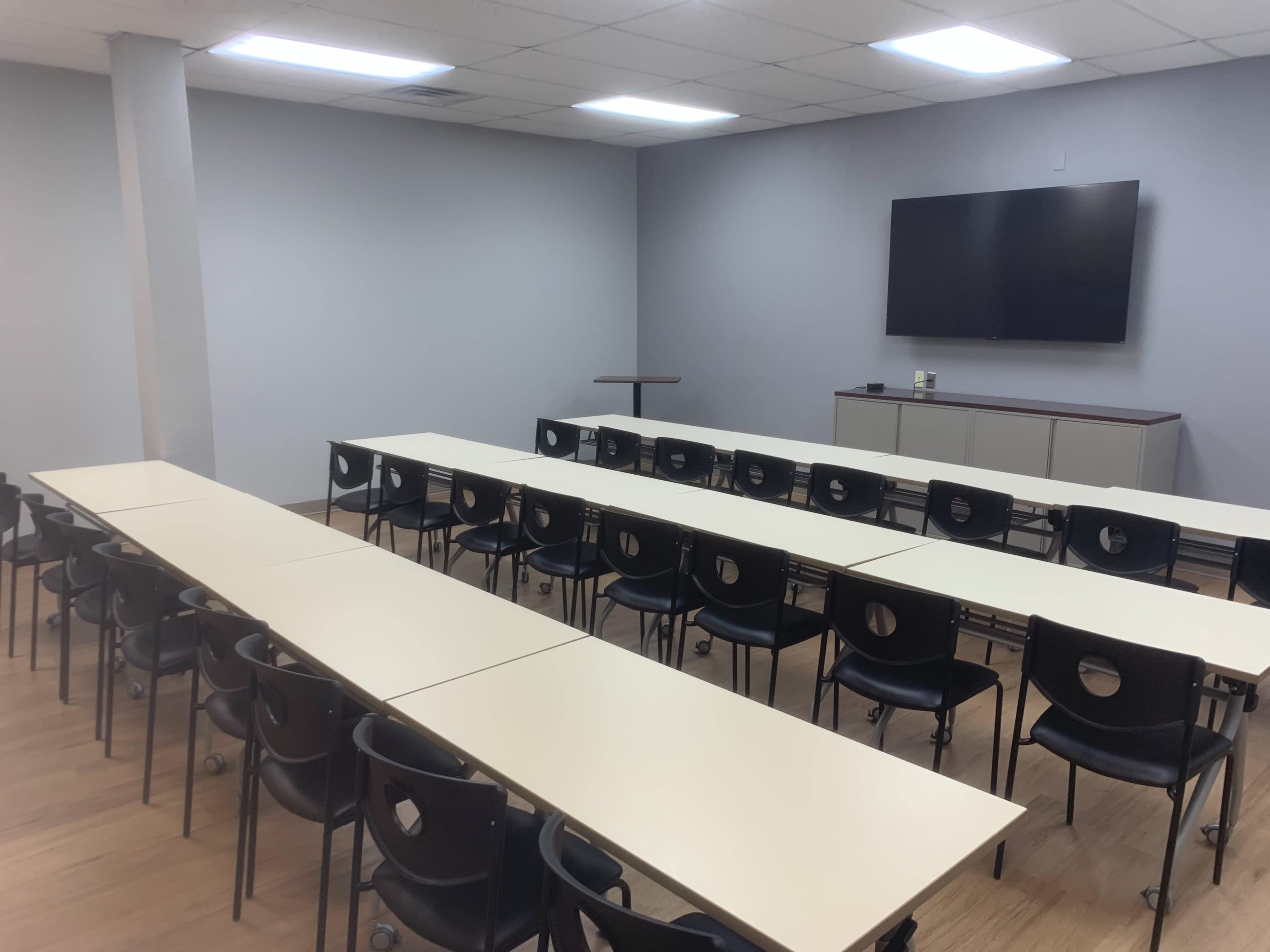 A classroom or meeting room with several rows of tables and black chairs facing a wall-mounted television.