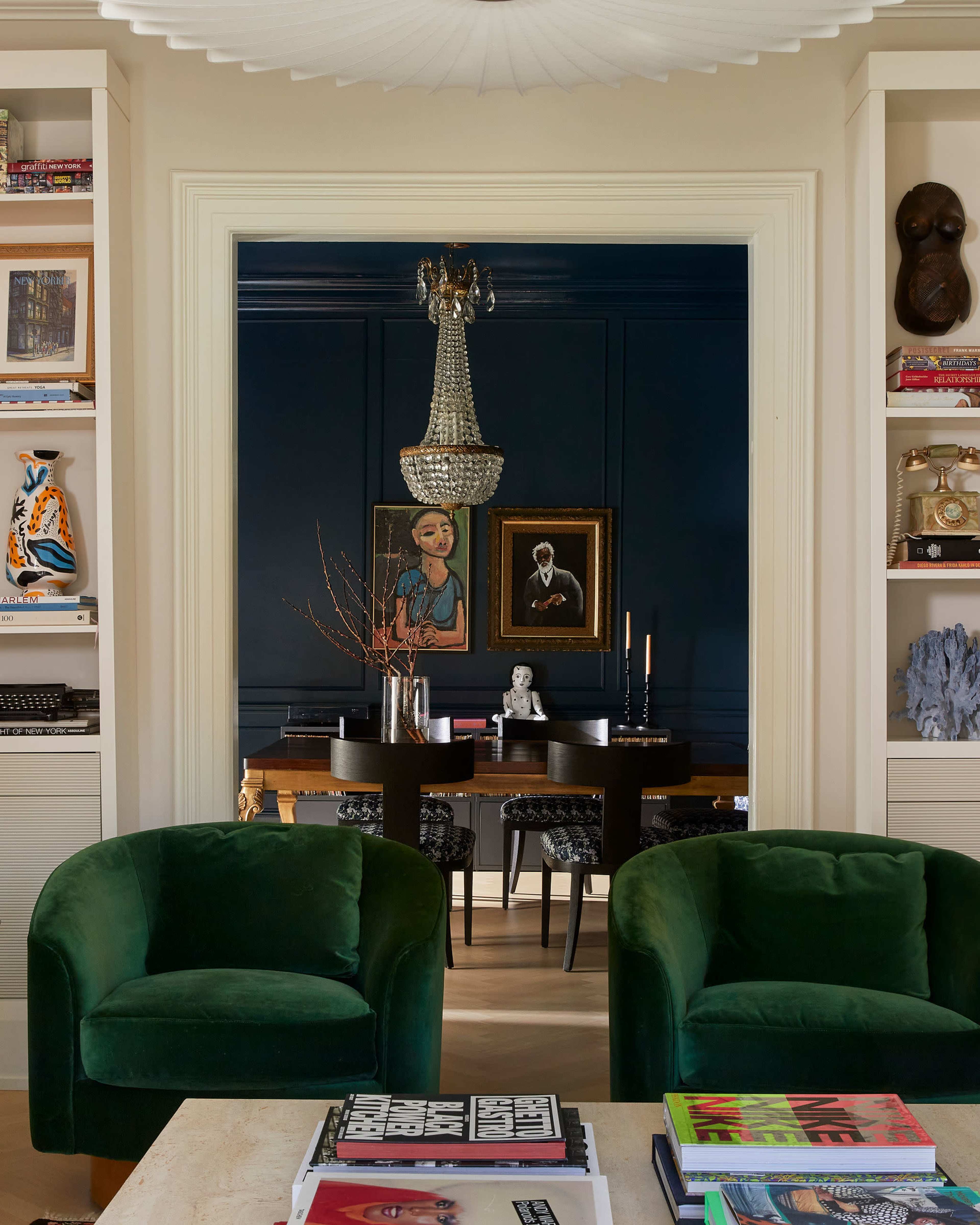 The image depicts a stylish living space featuring two green armchairs in the foreground, with a dining area visible through an archway in the background, illuminated by an ornate chandelier.