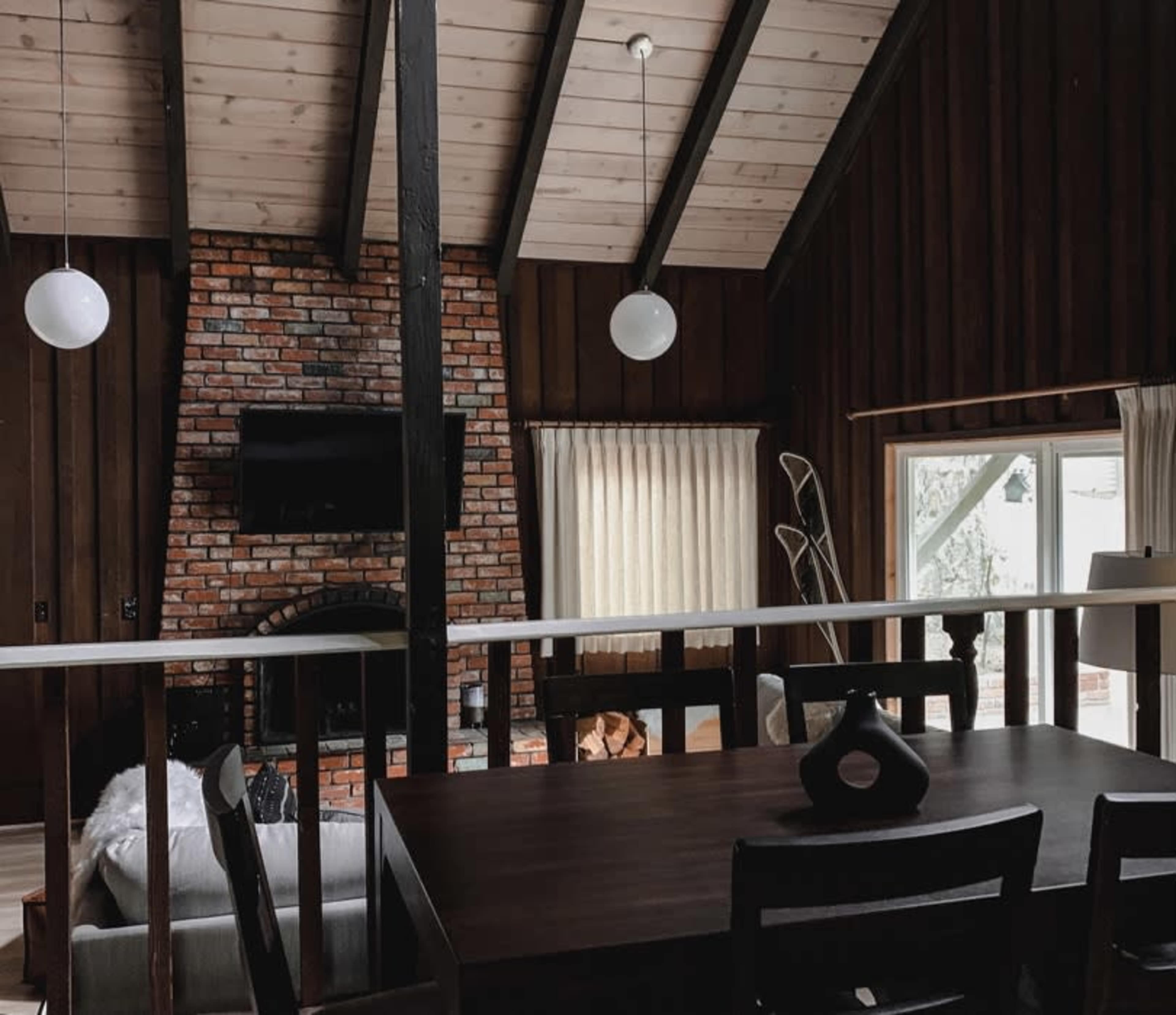 The interior of a cabin features a wooden dining table and a brick fireplace with a television above it, set against walls made of dark wood and a ceiling with exposed beams.