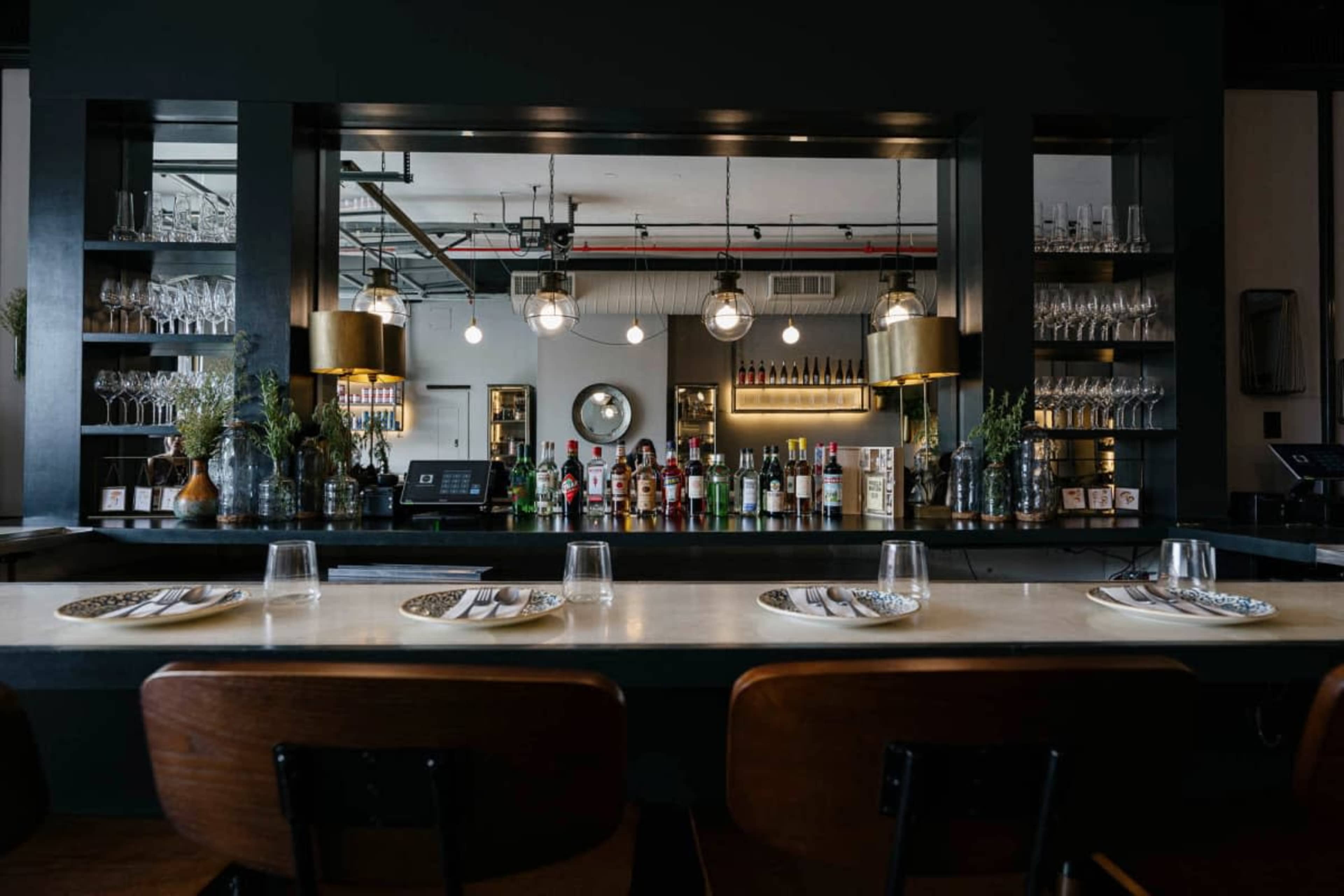 The image shows a modern bar area with a row of empty chairs, a drinks display, and a sleek counter adorned with dining plates and glassware.