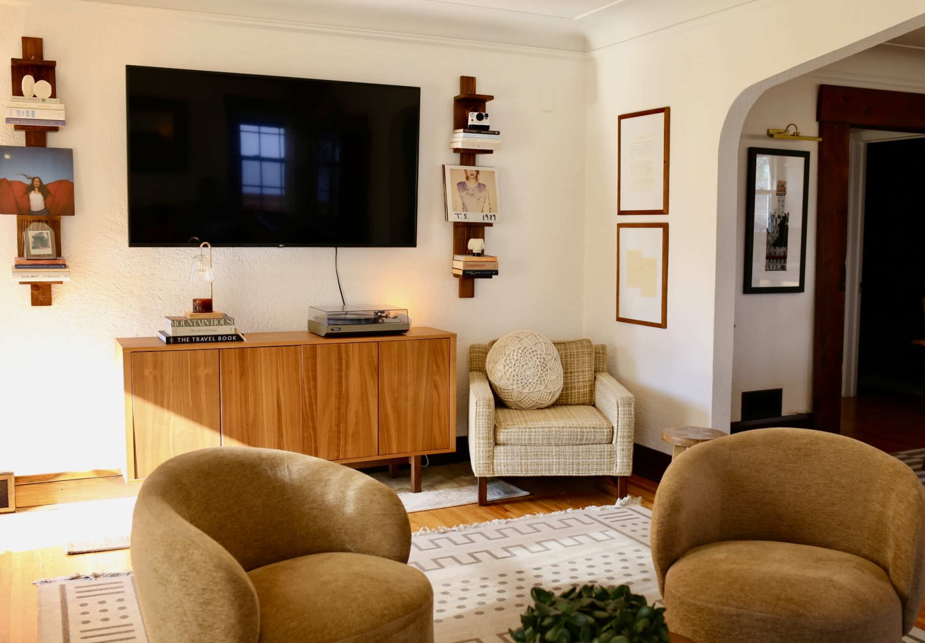 The image shows a modern living room featuring a television mounted on the wall, a wooden sideboard, and two upholstered chairs facing a patterned rug.
