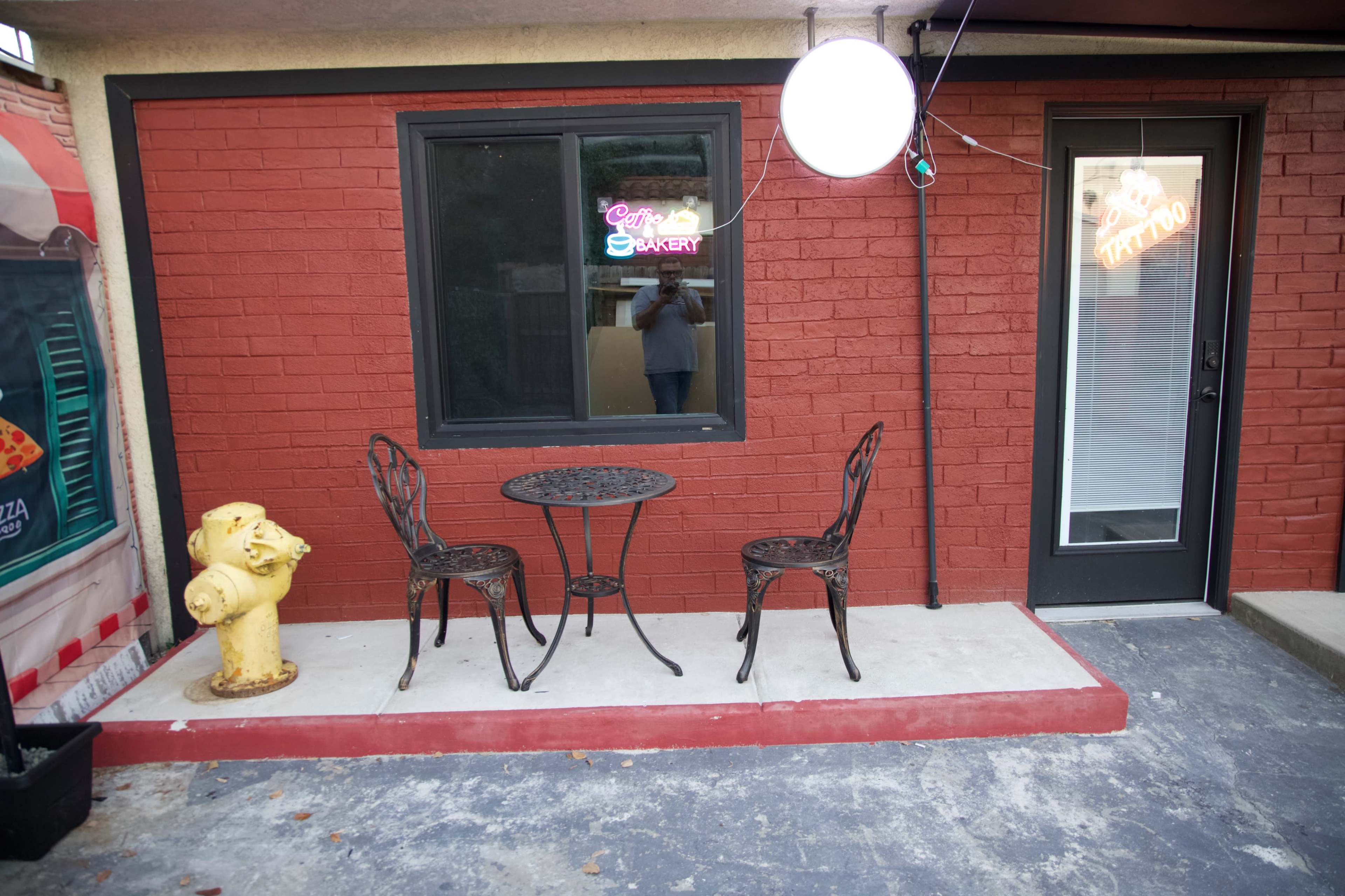A small outdoor seating area features a round table and two chairs beside a bright red wall, with a fire hydrant nearby and a neon sign in the window.