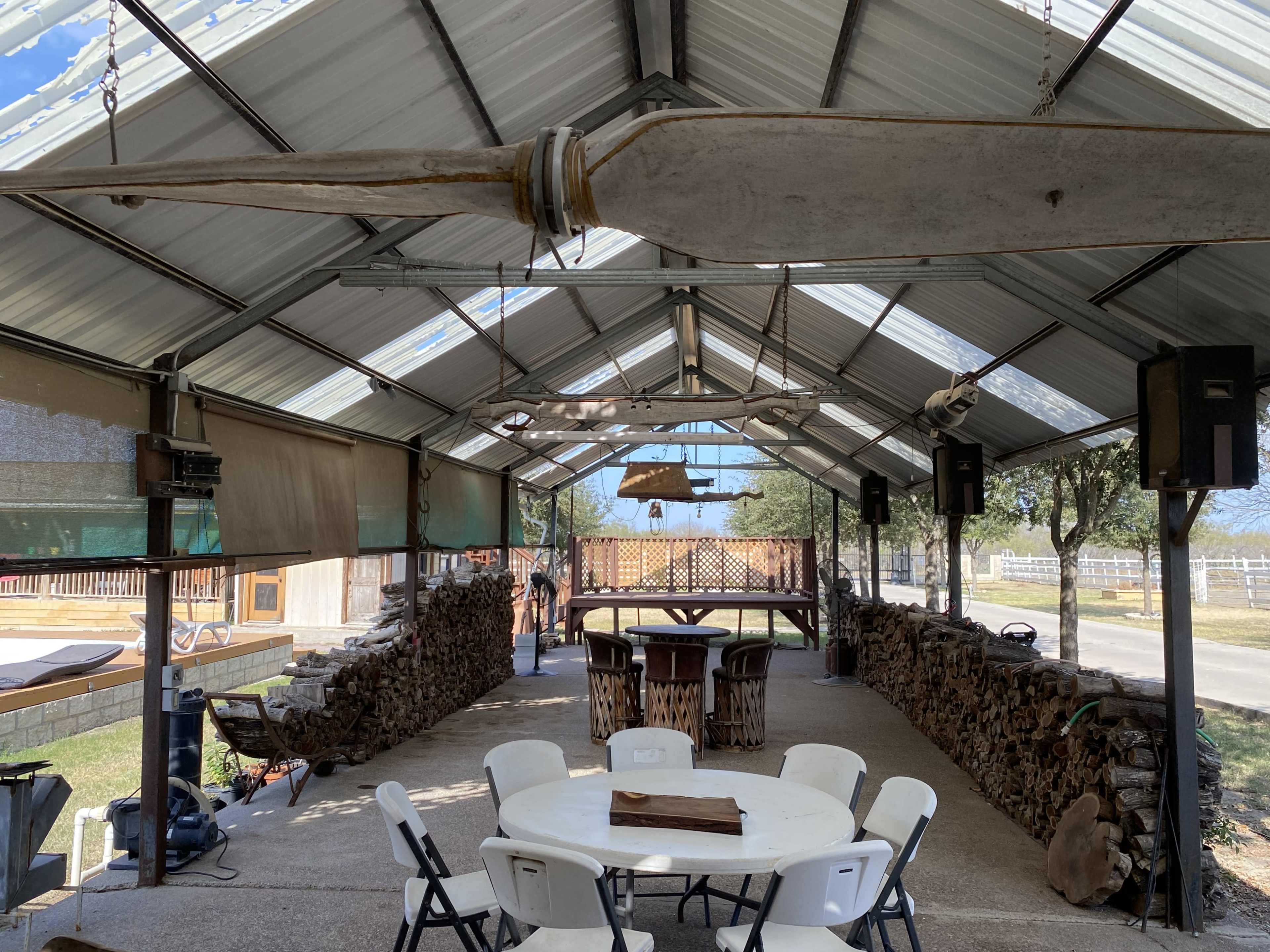 The image shows a large, open pavilion with a high metal roof, featuring a central circular table surrounded by folding chairs, and walls lined with stacked firewood.