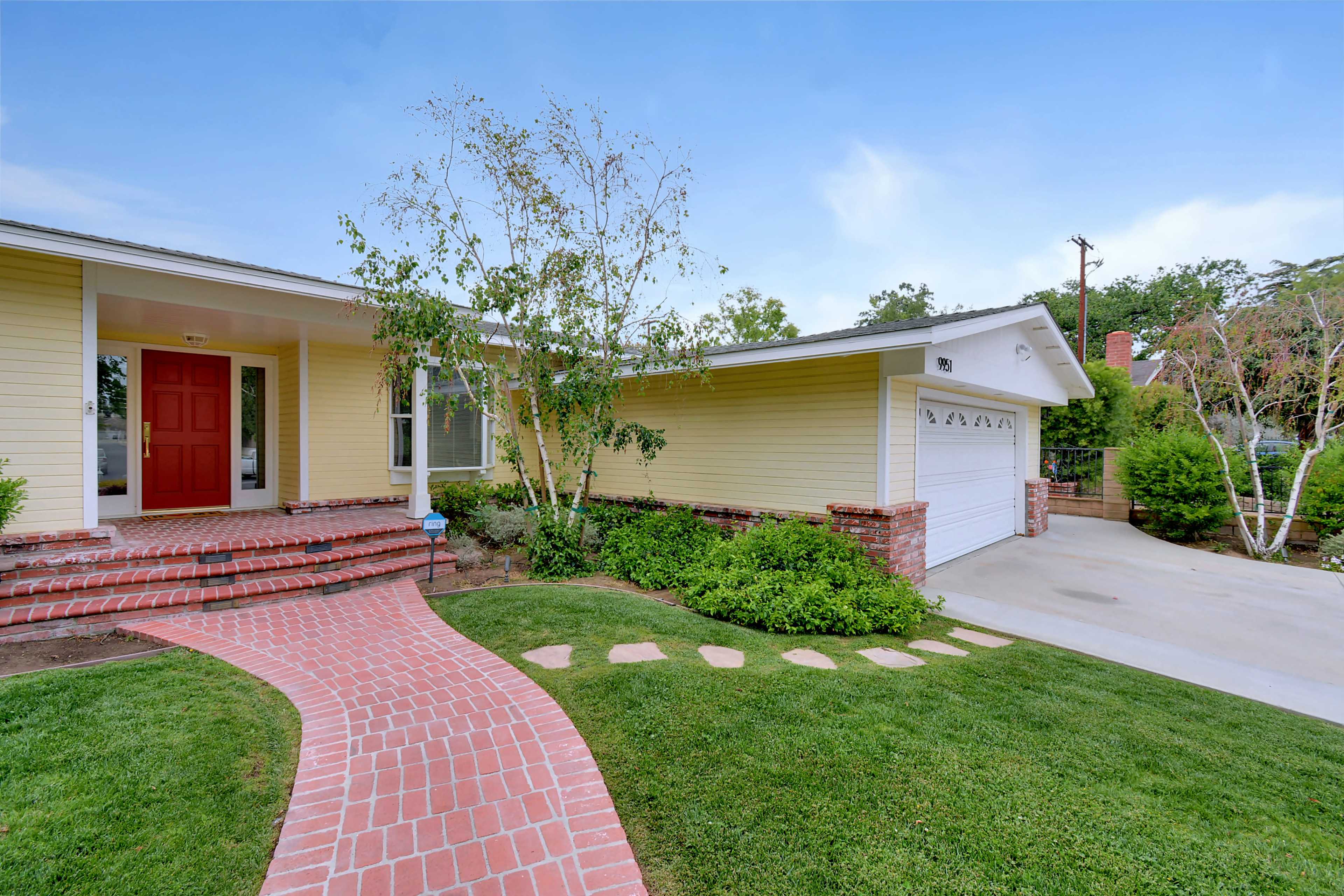 The image shows a single-story house with a red front door, a brick pathway, and a well-maintained lawn.