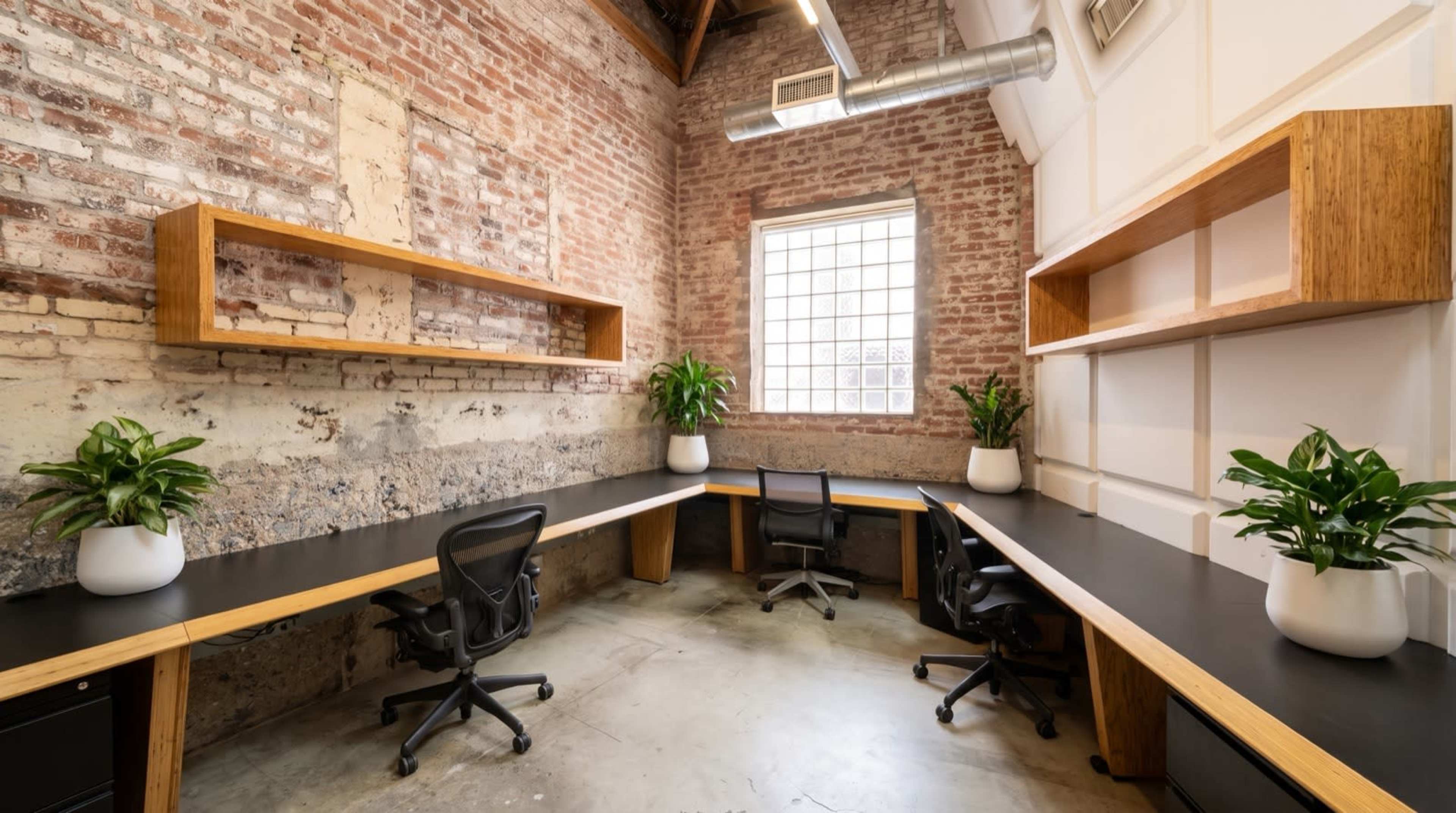 The image depicts a modern office space featuring a corner layout with two black desks, four ergonomic chairs, and two potted plants on each desk against a brick wall.