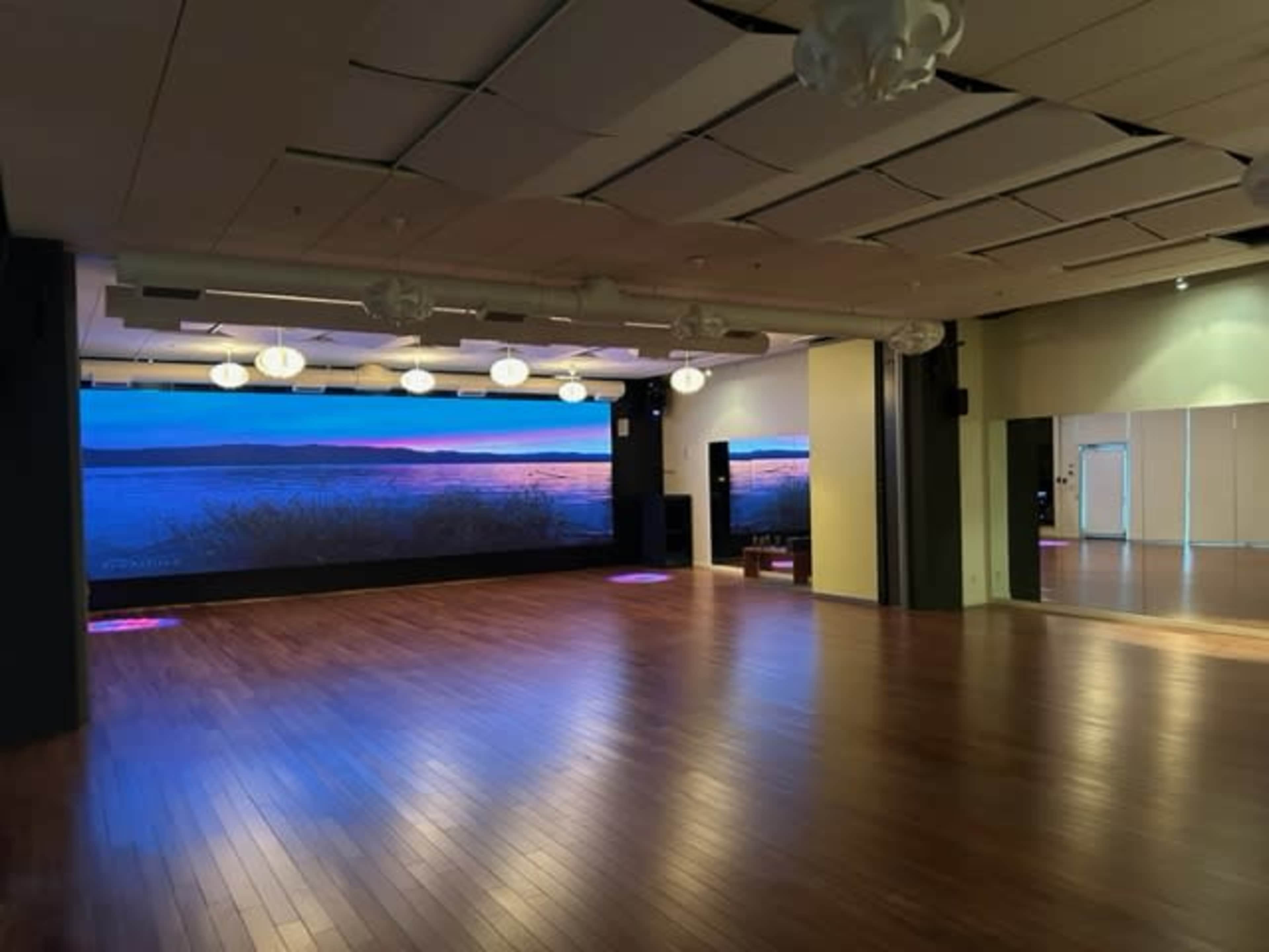A spacious studio with a polished wooden floor, large screens displaying a scenic view, and soft lighting fixtures on the ceiling.