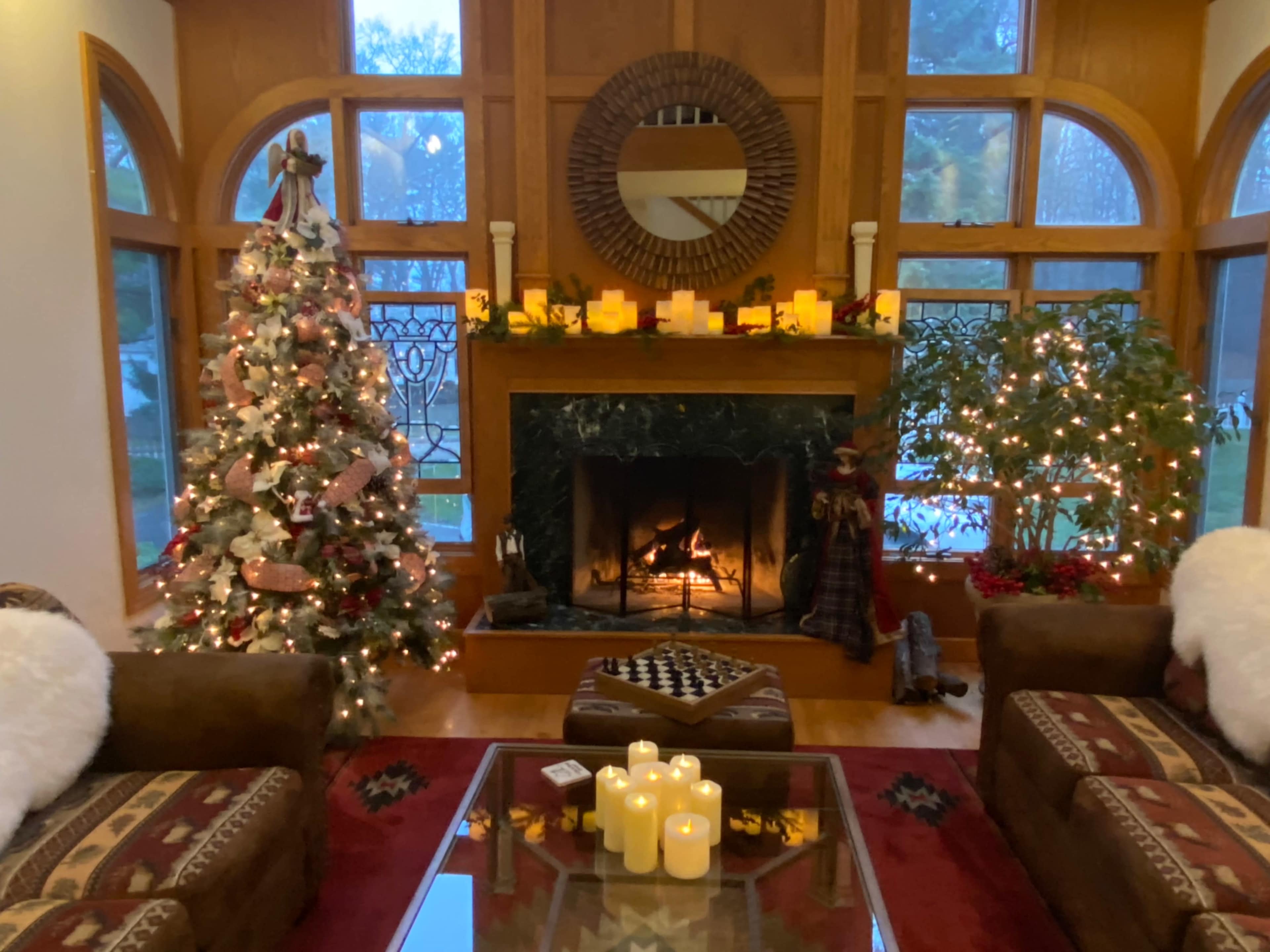 A cozy living room features a decorated Christmas tree, a fireplace with a visible fire, and warm lighting from candles on a coffee table.