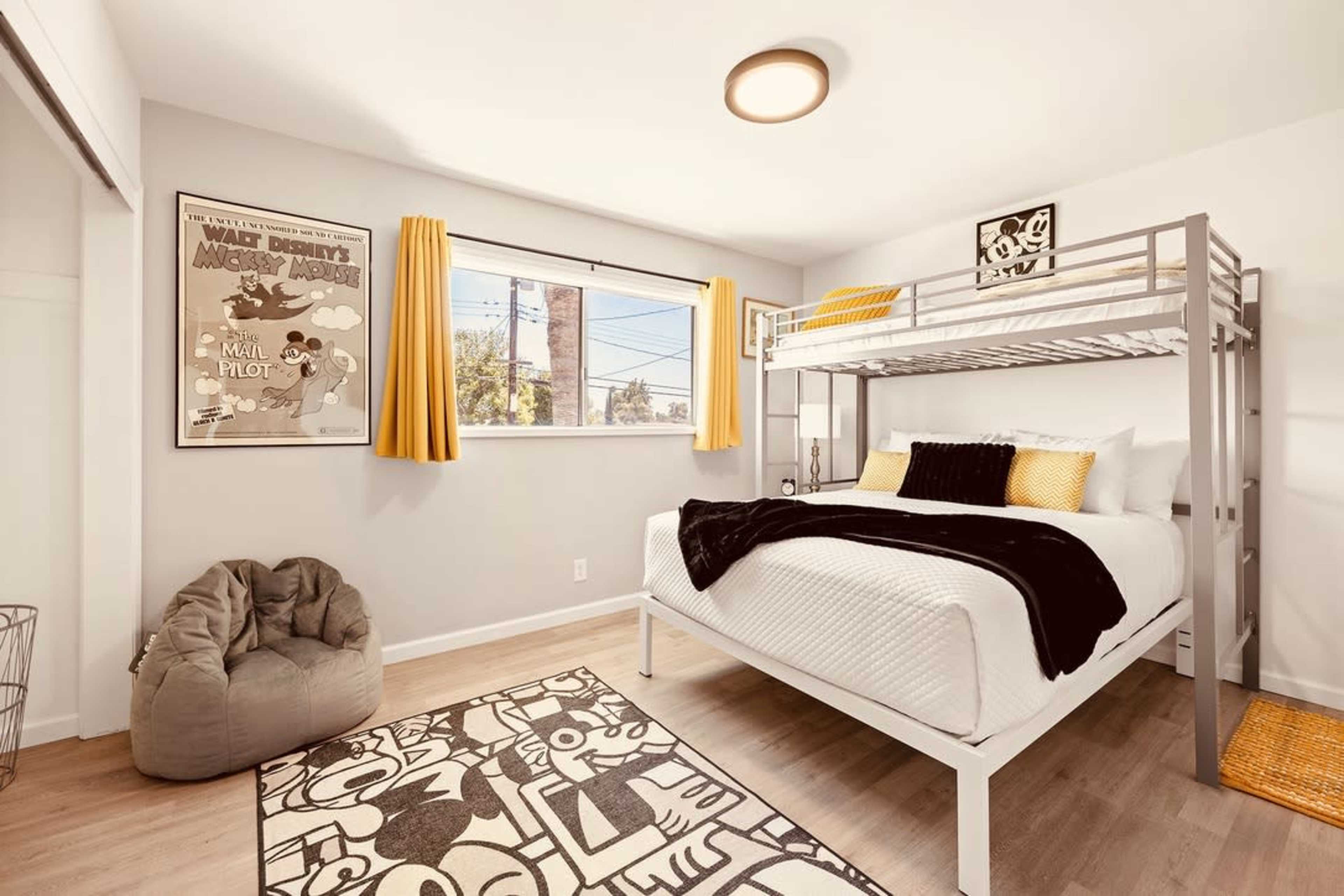A well-lit bedroom with a lofted bed, yellow curtains, a bean bag chair, and a rug featuring a graphic design.