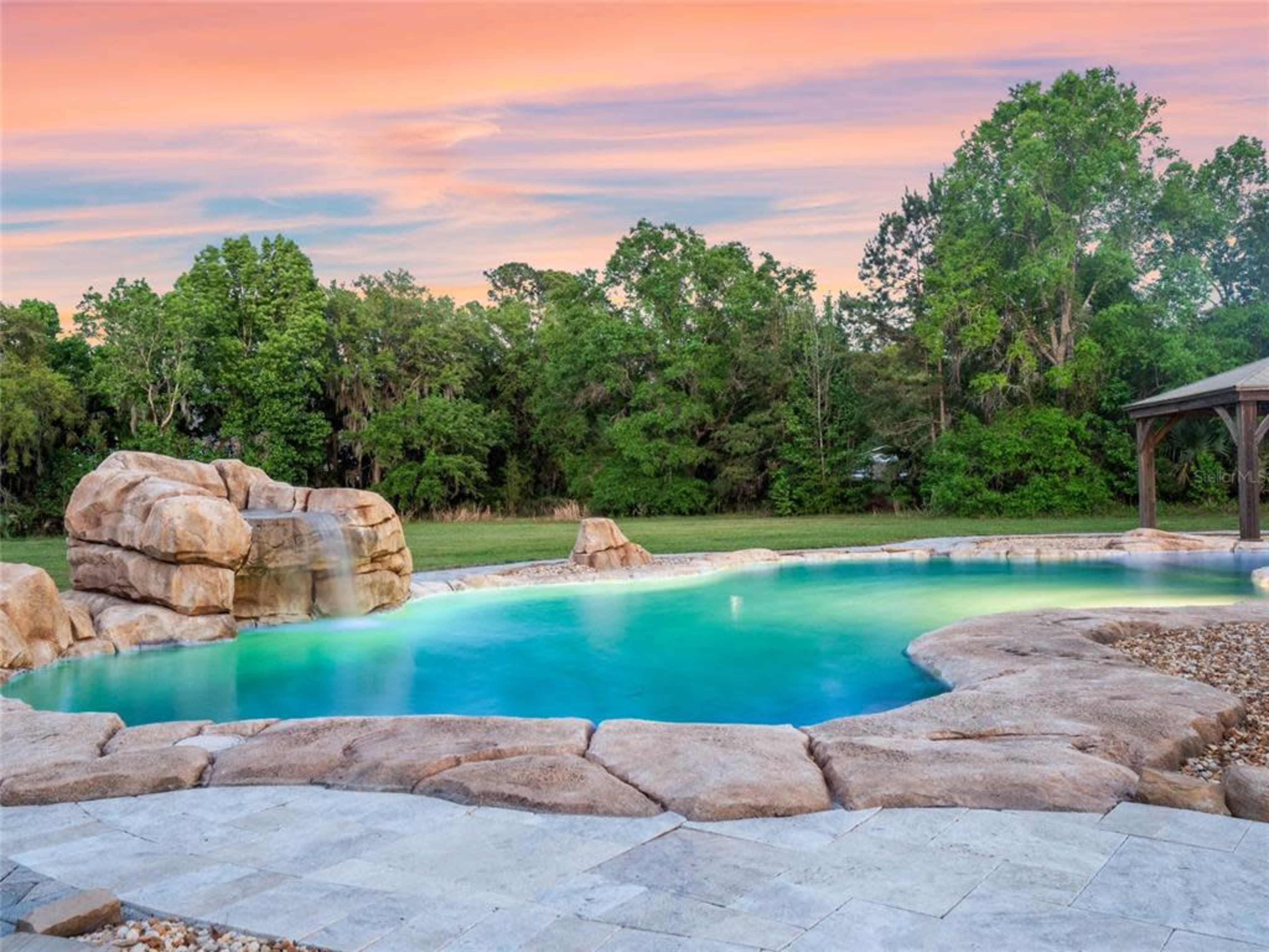 A large, natural-looking swimming pool is surrounded by rock formations and lush greenery under a colorful sky at sunset.