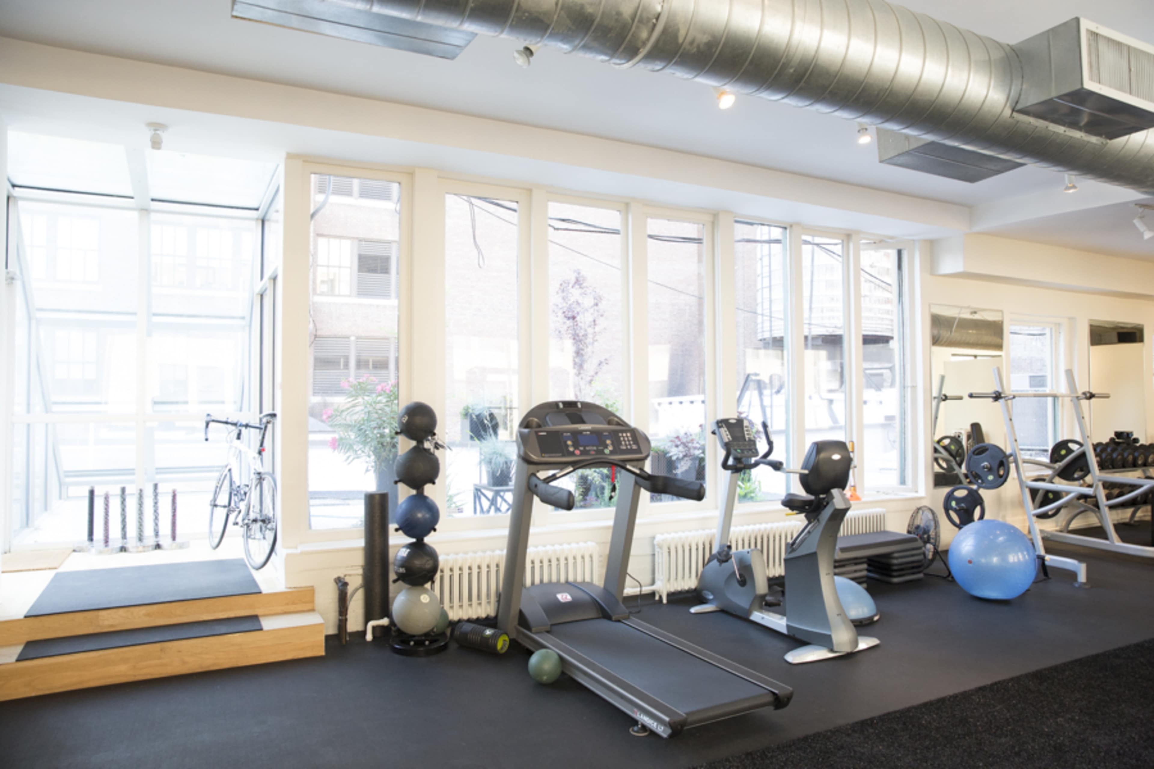 A fitness room features a treadmill, stationary bike, exercise balls, and free weights, with large windows allowing natural light to enter.