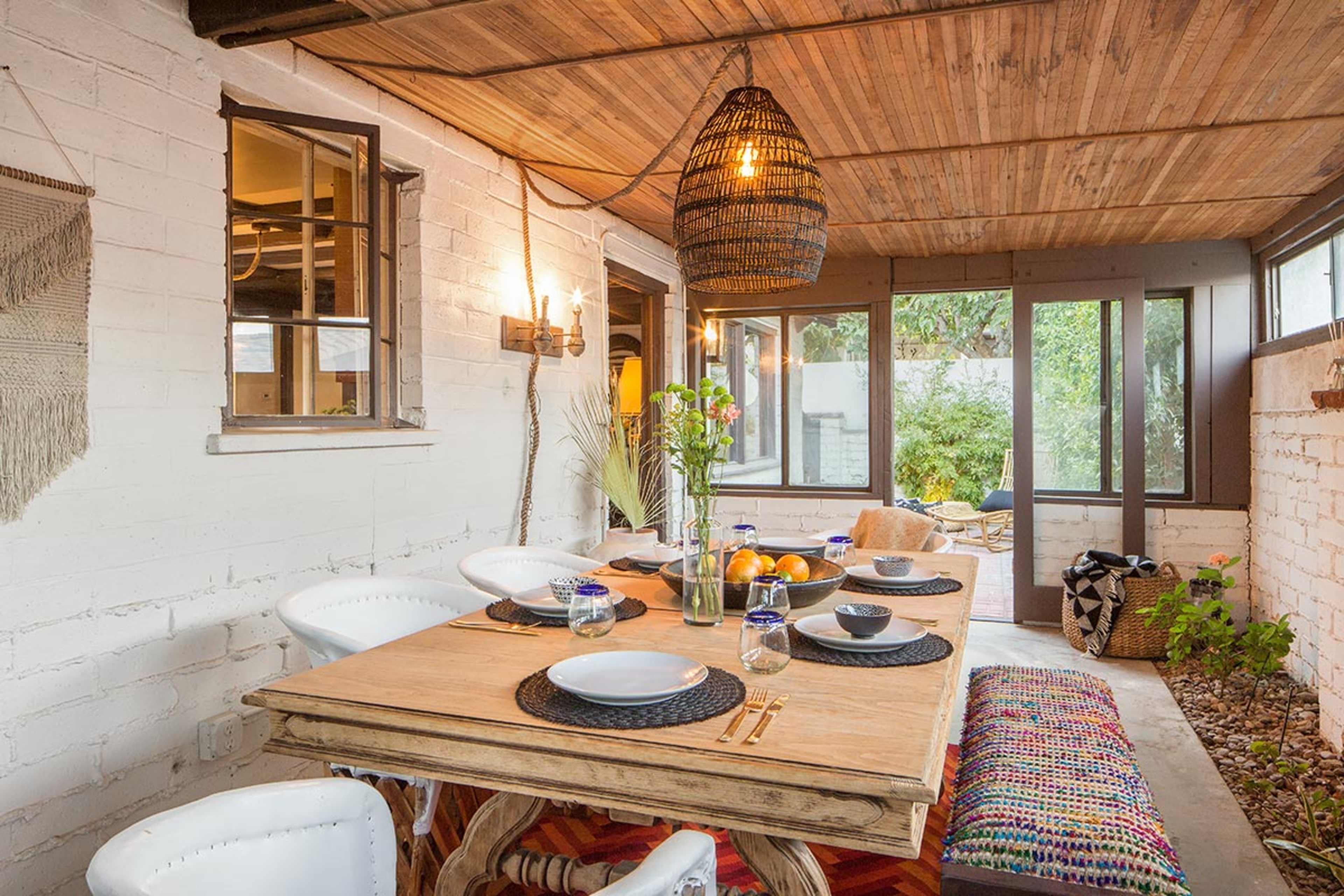 The image shows a sunlit indoor dining area with a wooden table set for a meal, surrounded by white chairs and decorated with plants and a woven light fixture.
