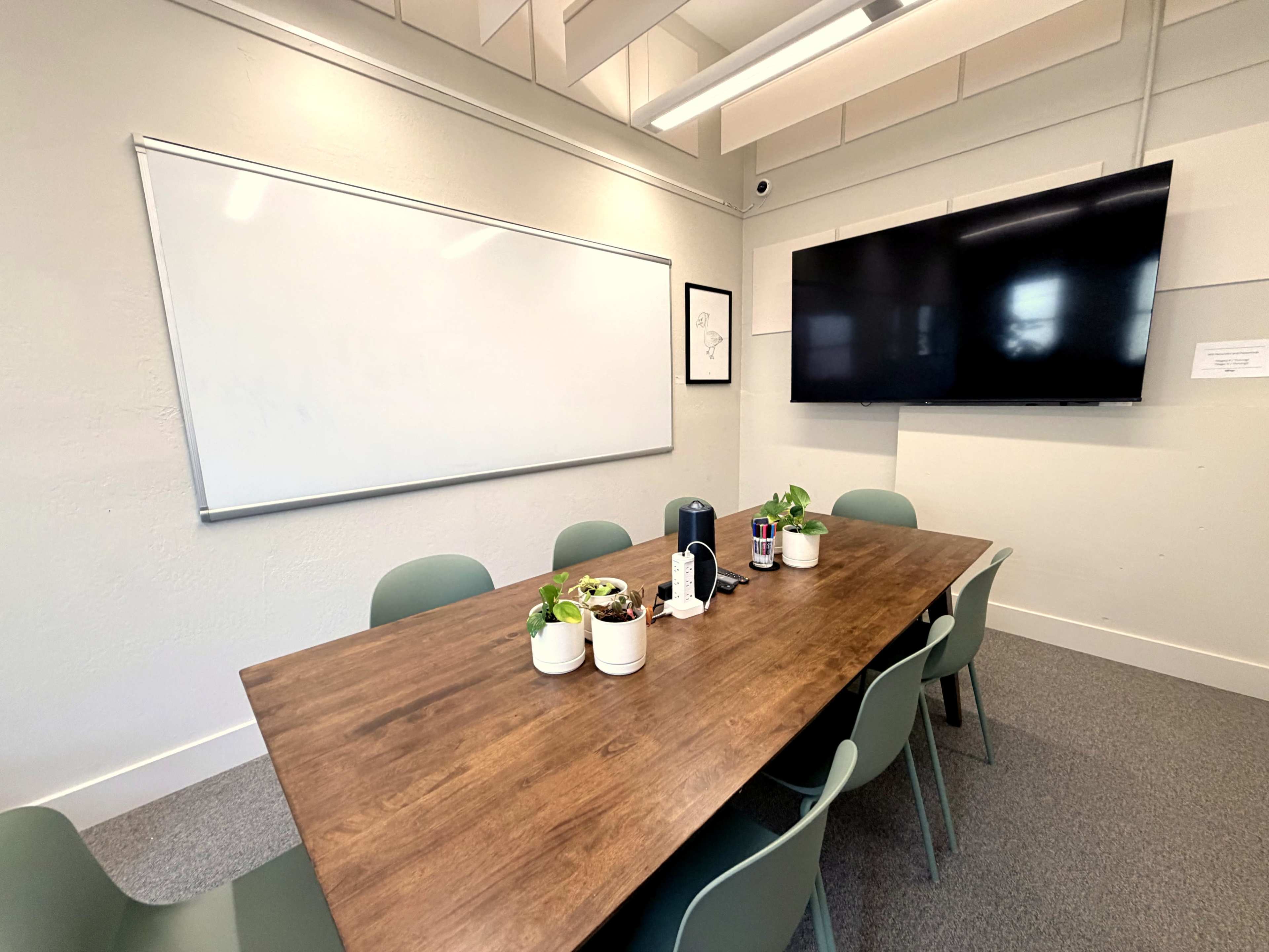 The image shows a conference room with a large wooden table surrounded by green chairs, a whiteboard on one wall, and a television screen mounted on another wall.
