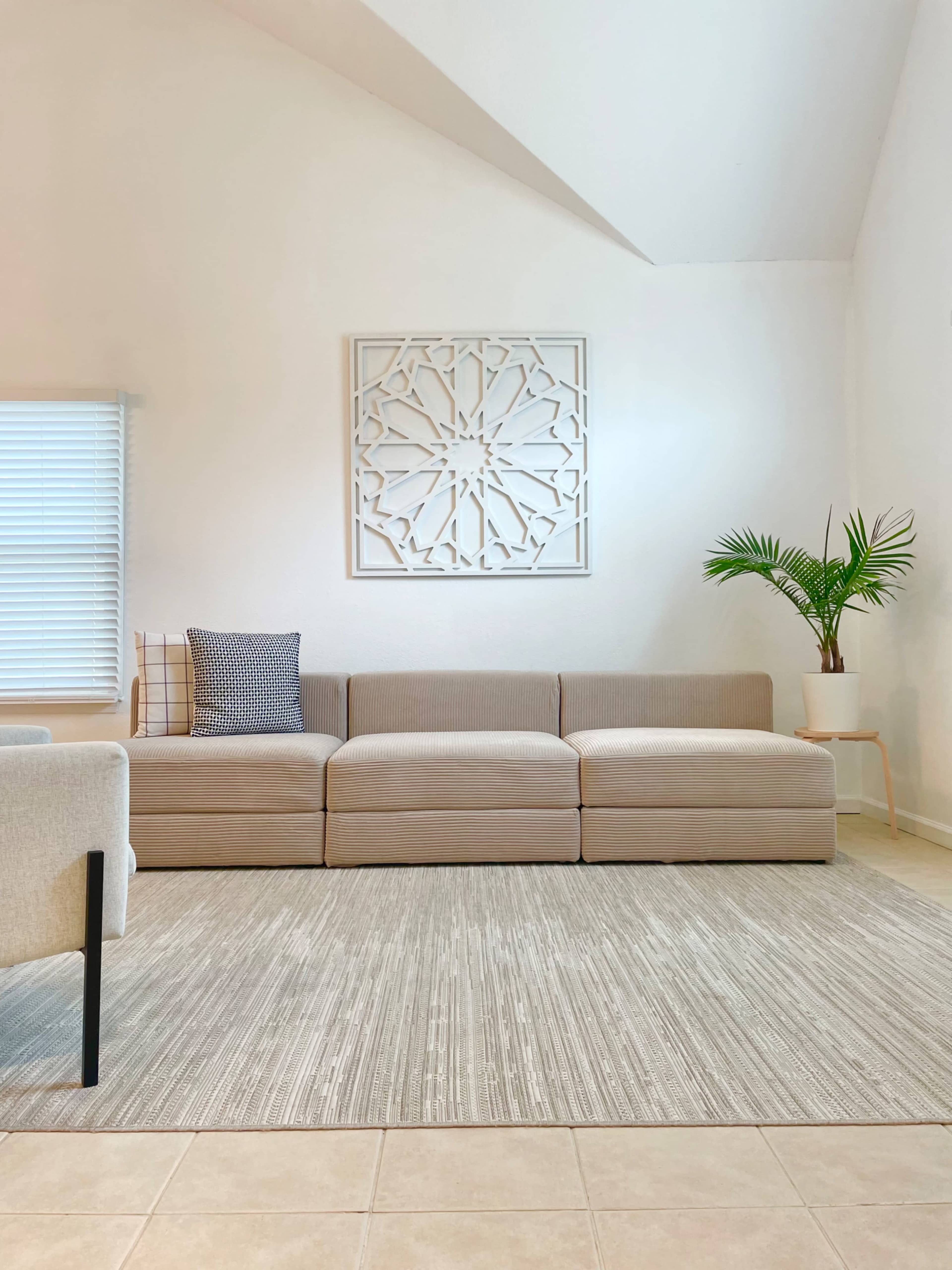 A light-colored living room with a sectional sofa, a decorative wall panel, a plant, and a neutral rug on tiled flooring.