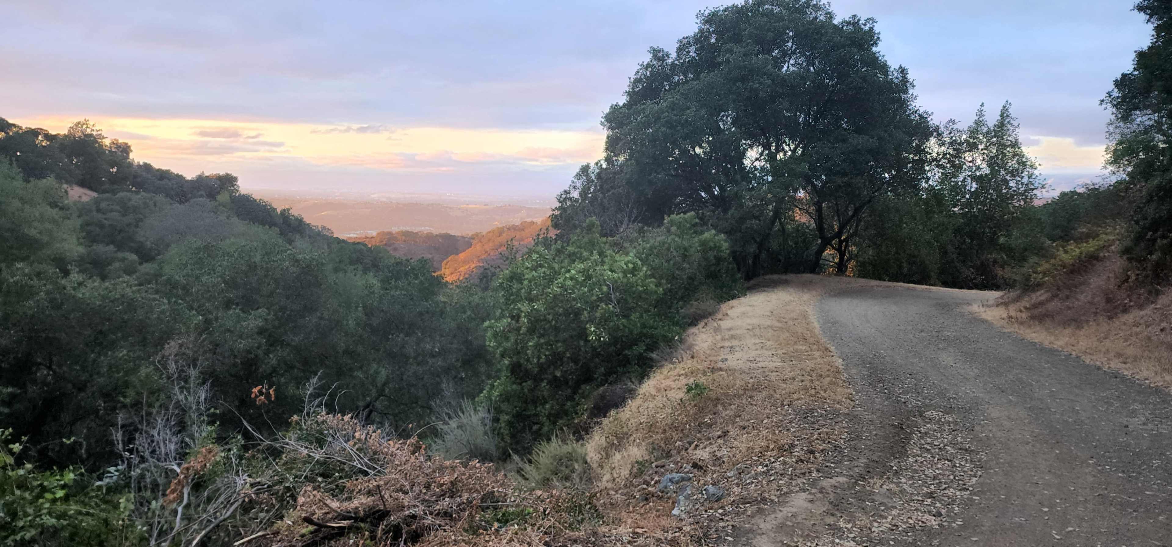 A winding dirt path leads through green trees, with a view of rolling hills and a colorful sunset in the distance.
