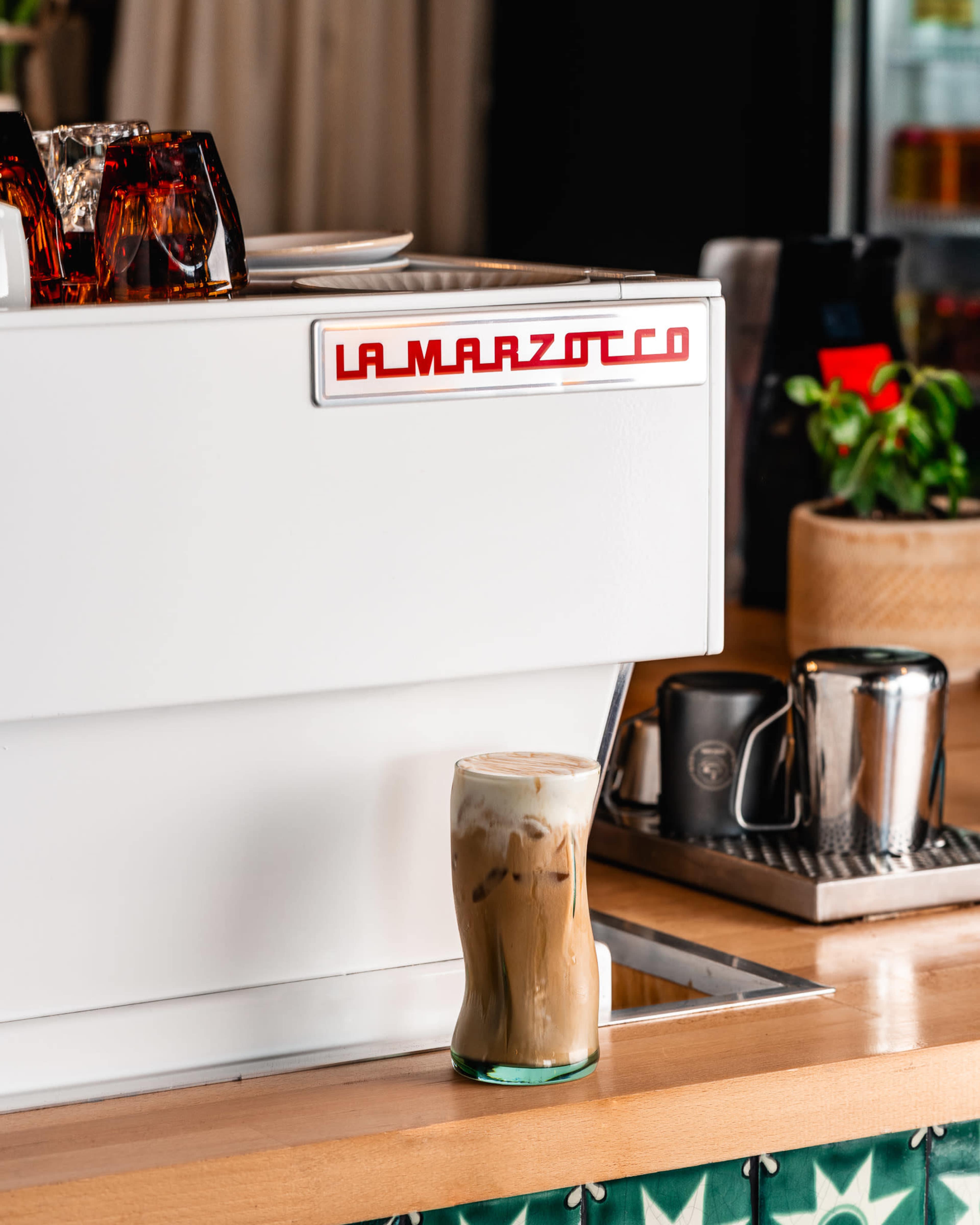A glass of iced coffee is positioned in front of a La Marzocco espresso machine on a wooden countertop.