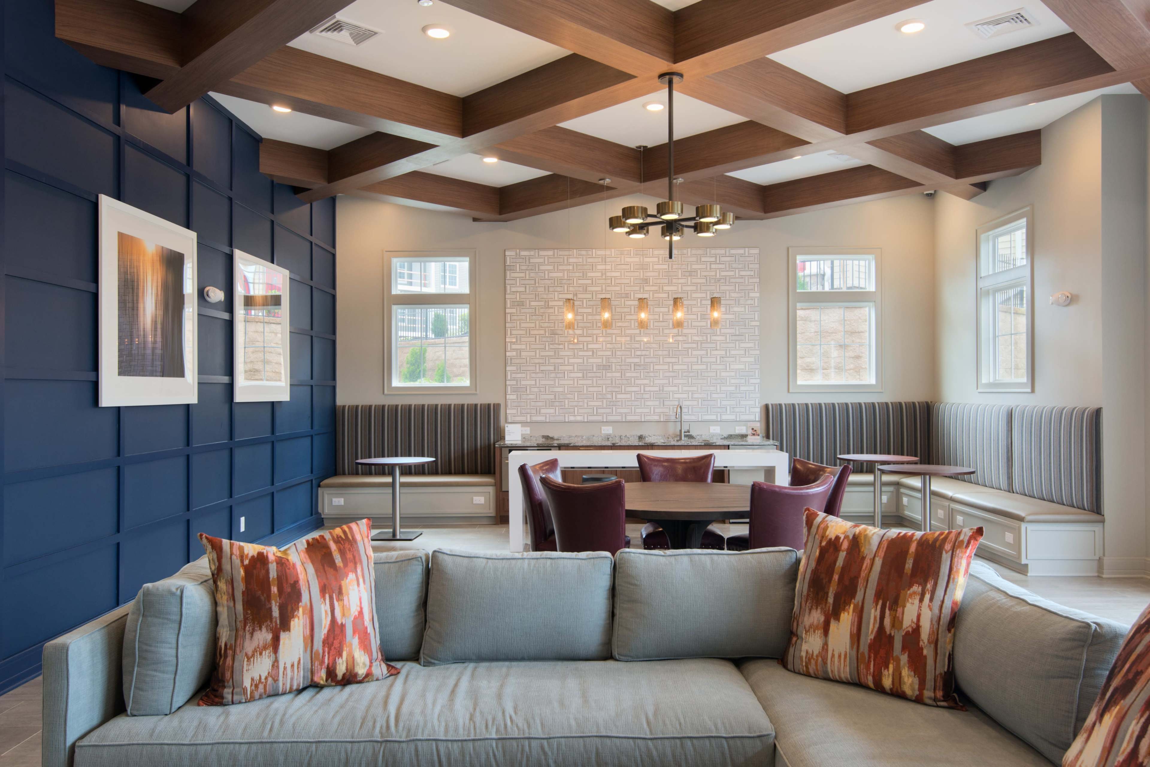 A modern lounge area featuring a gray sofa with colorful pillows, a coffered wooden ceiling, and a dining nook with tables and benches against a light brick wall.