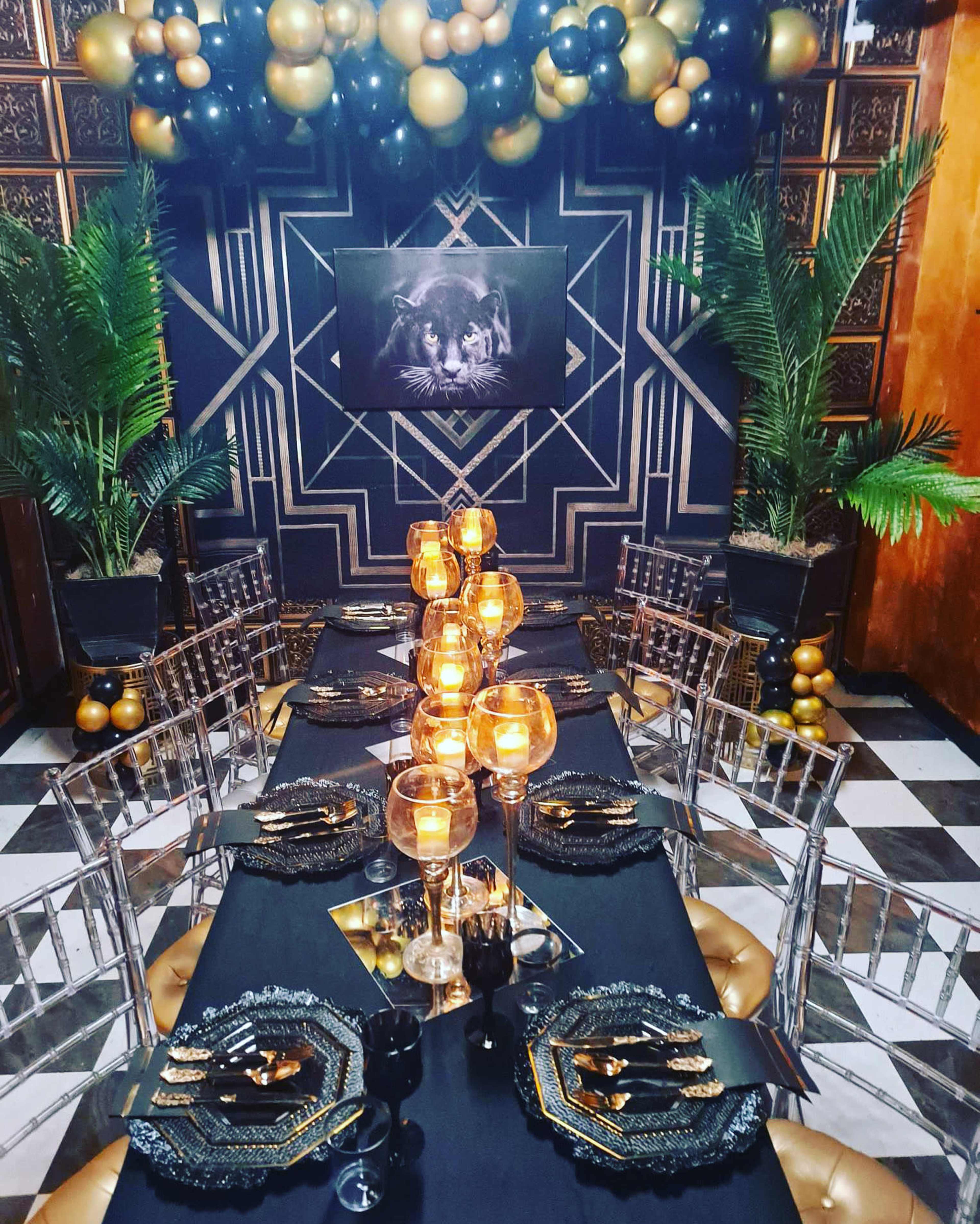A decorated dining area with a long black table set with golden and black tableware, illuminated by candlelight, surrounded by lush greenery and a backdrop of balloons in gold, black, and clear colors.