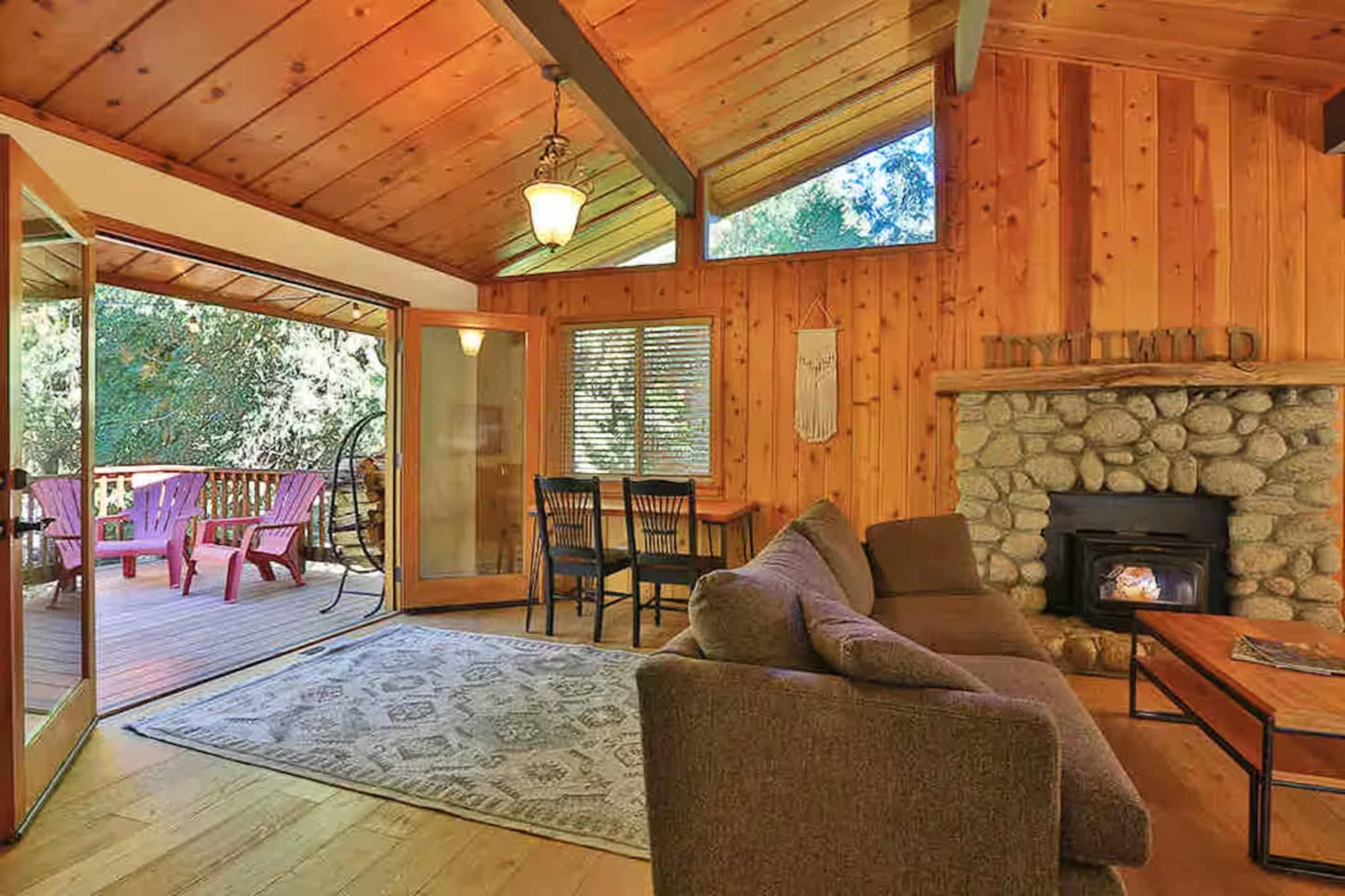 The image shows a cozy wooden cabin interior featuring a stone fireplace, a comfortable sofa, and a view of an outdoor seating area through large glass doors.