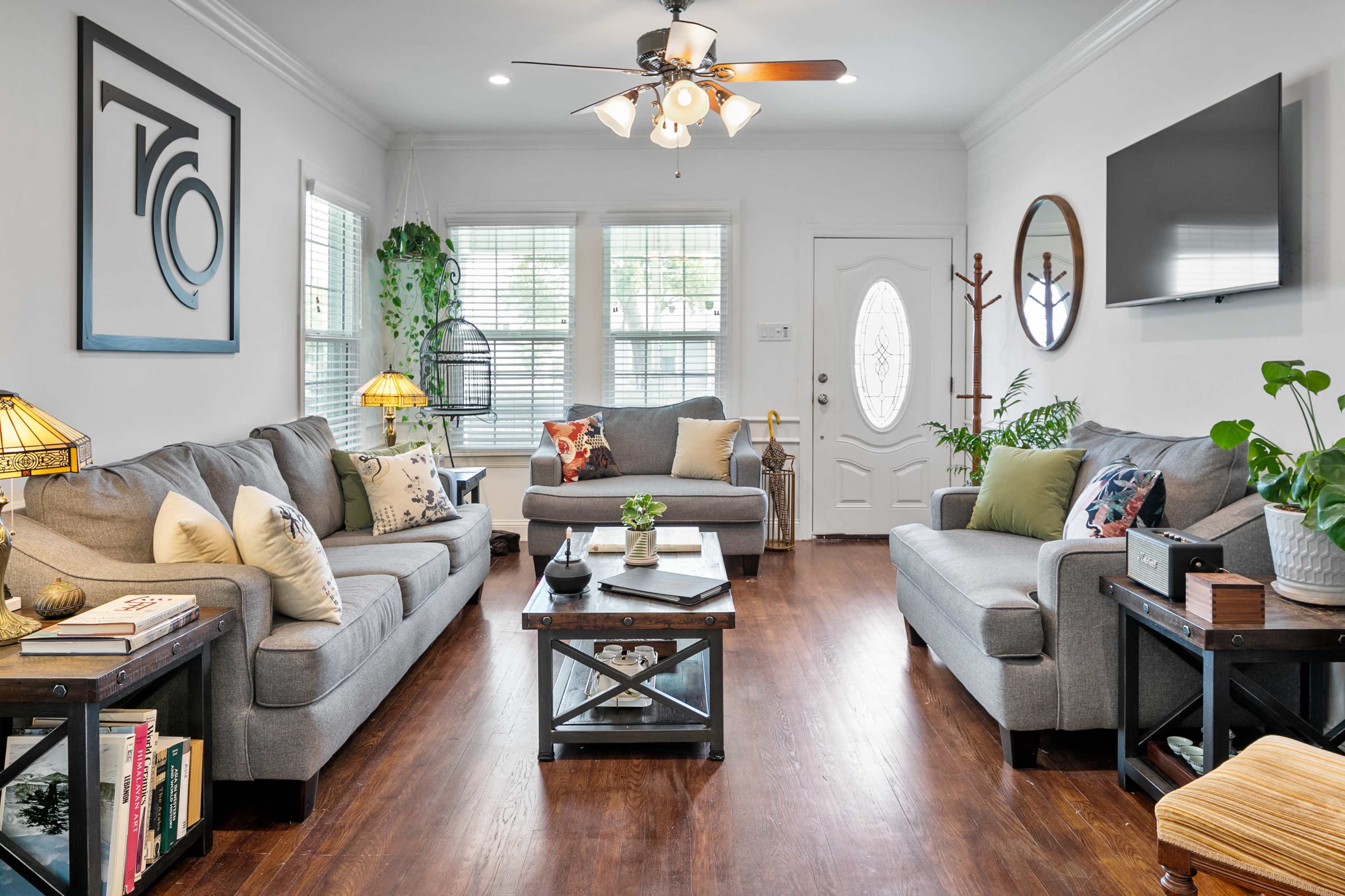 The living room features two gray sofas, a coffee table, potted plants, and a large window with natural light.