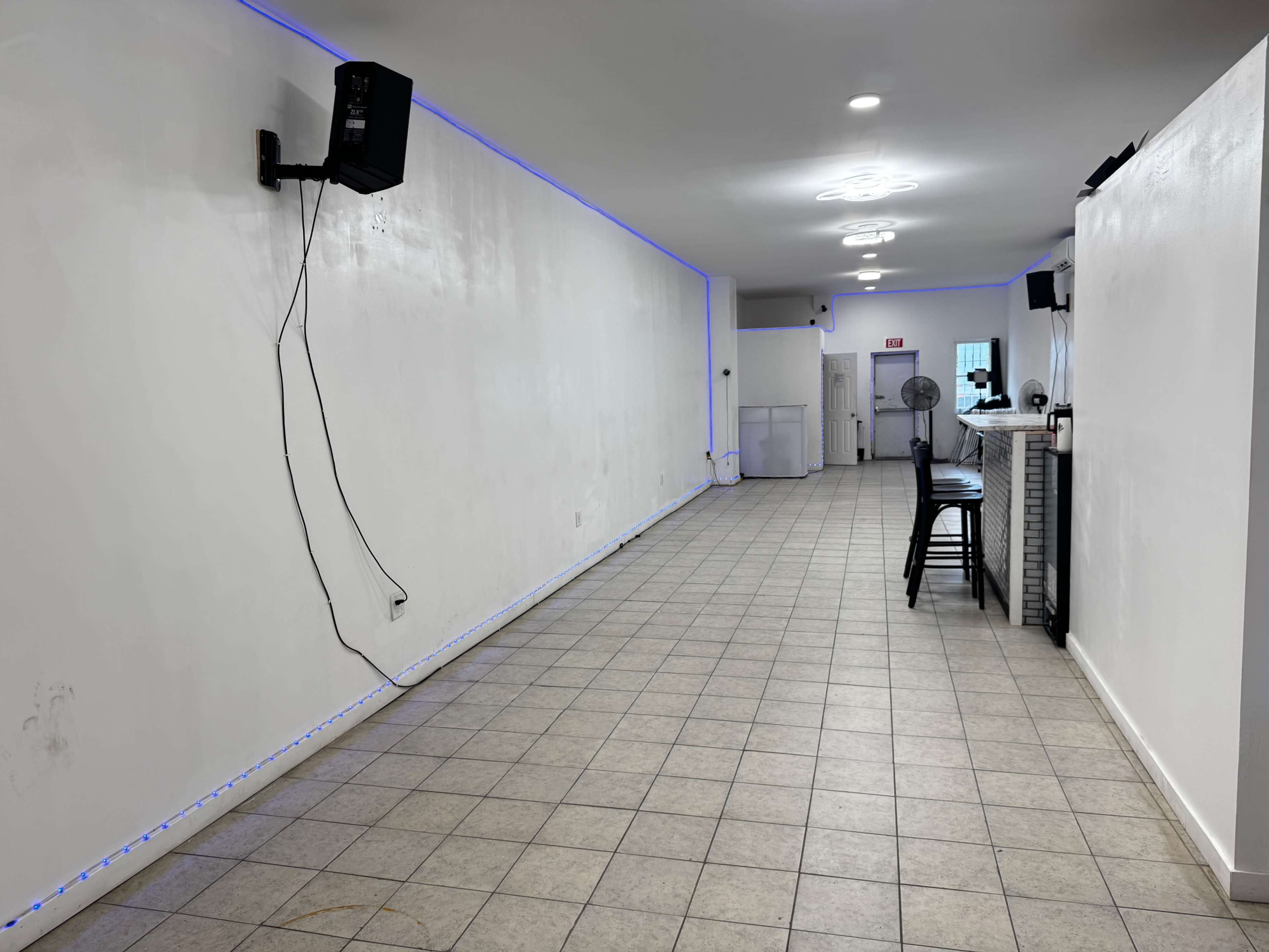 The image shows an empty indoor space with white walls, tiled flooring, and blue LED lighting along the base, featuring a bar area and a fan on one side.