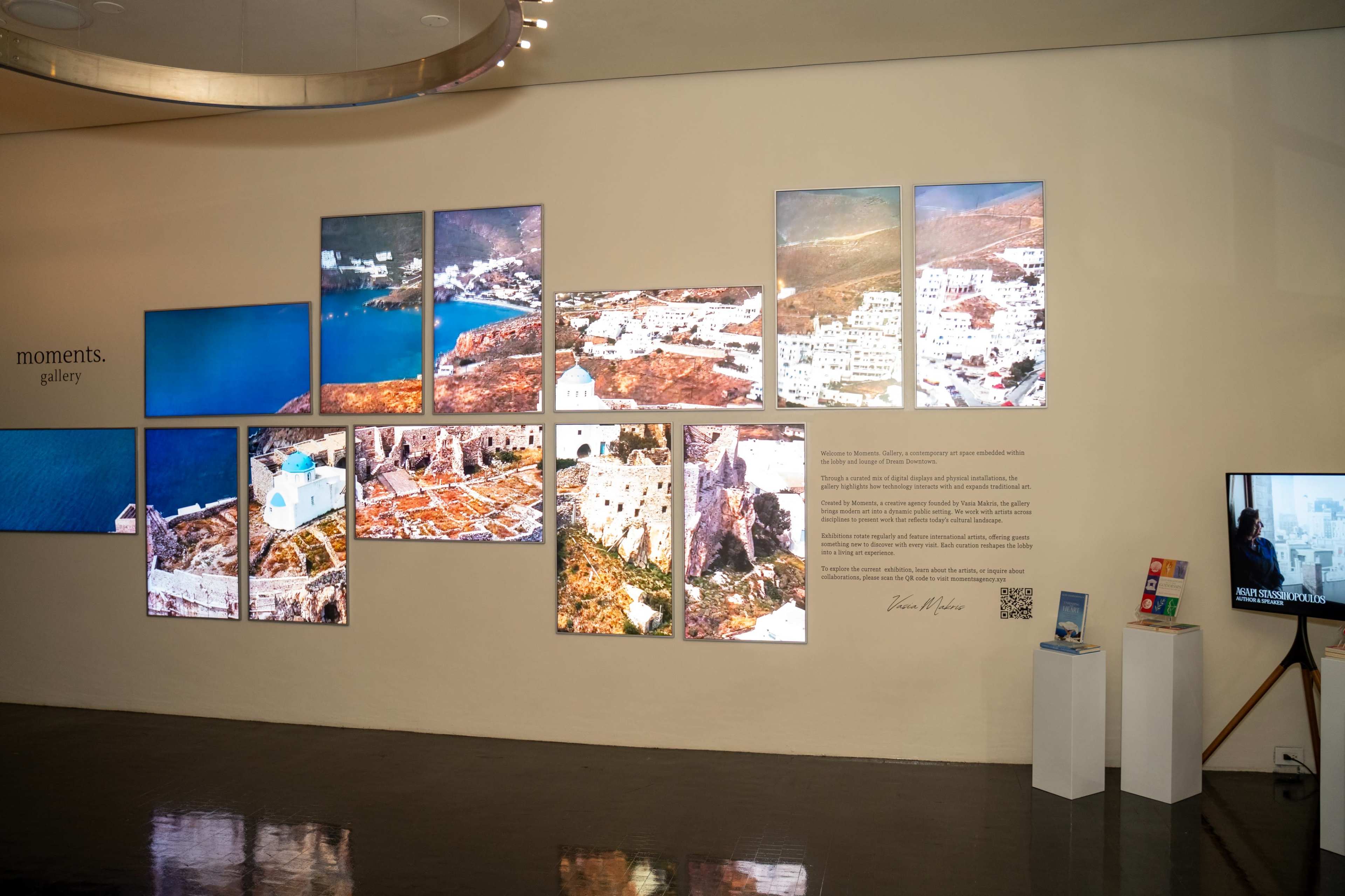 The image shows a gallery wall displaying a series of framed photographs depicting scenic views of a coastal village along with a small informational panel.