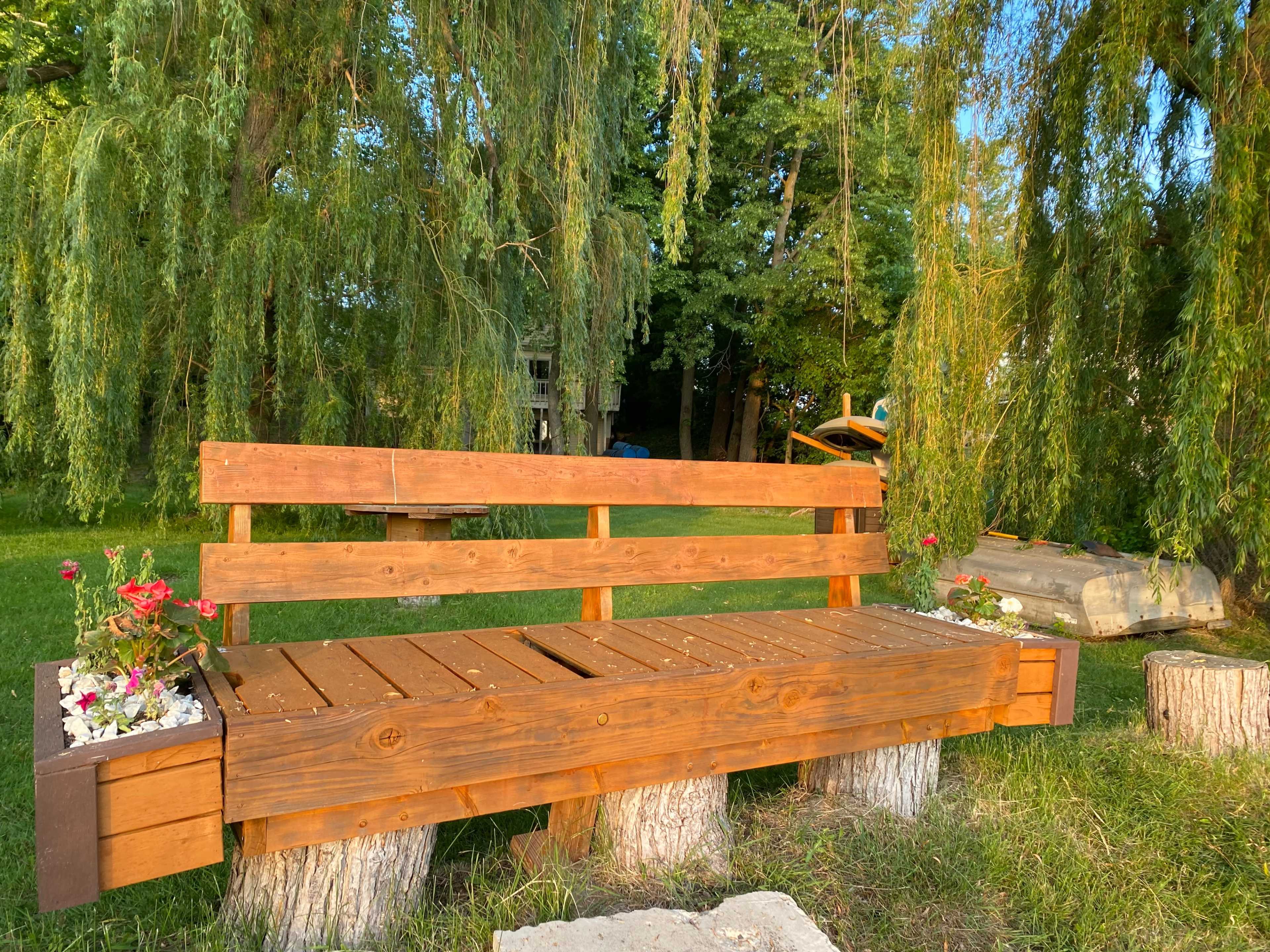 A wooden bench with flower planters is situated on tree stumps beneath a leafy canopy.