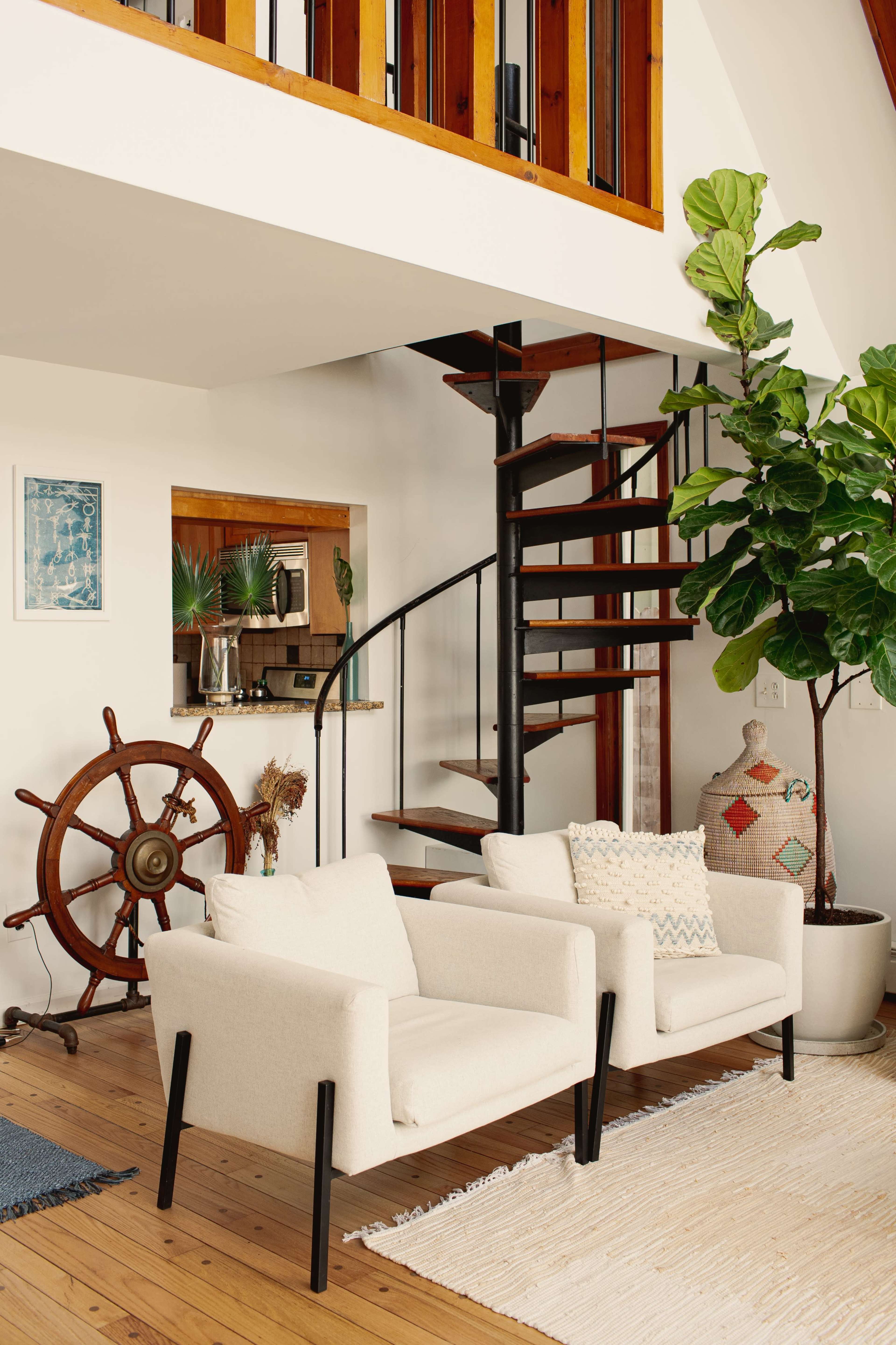 A bright living area features two cream-colored chairs, a spiral staircase, a potted plant, and a decorative ship's wheel.