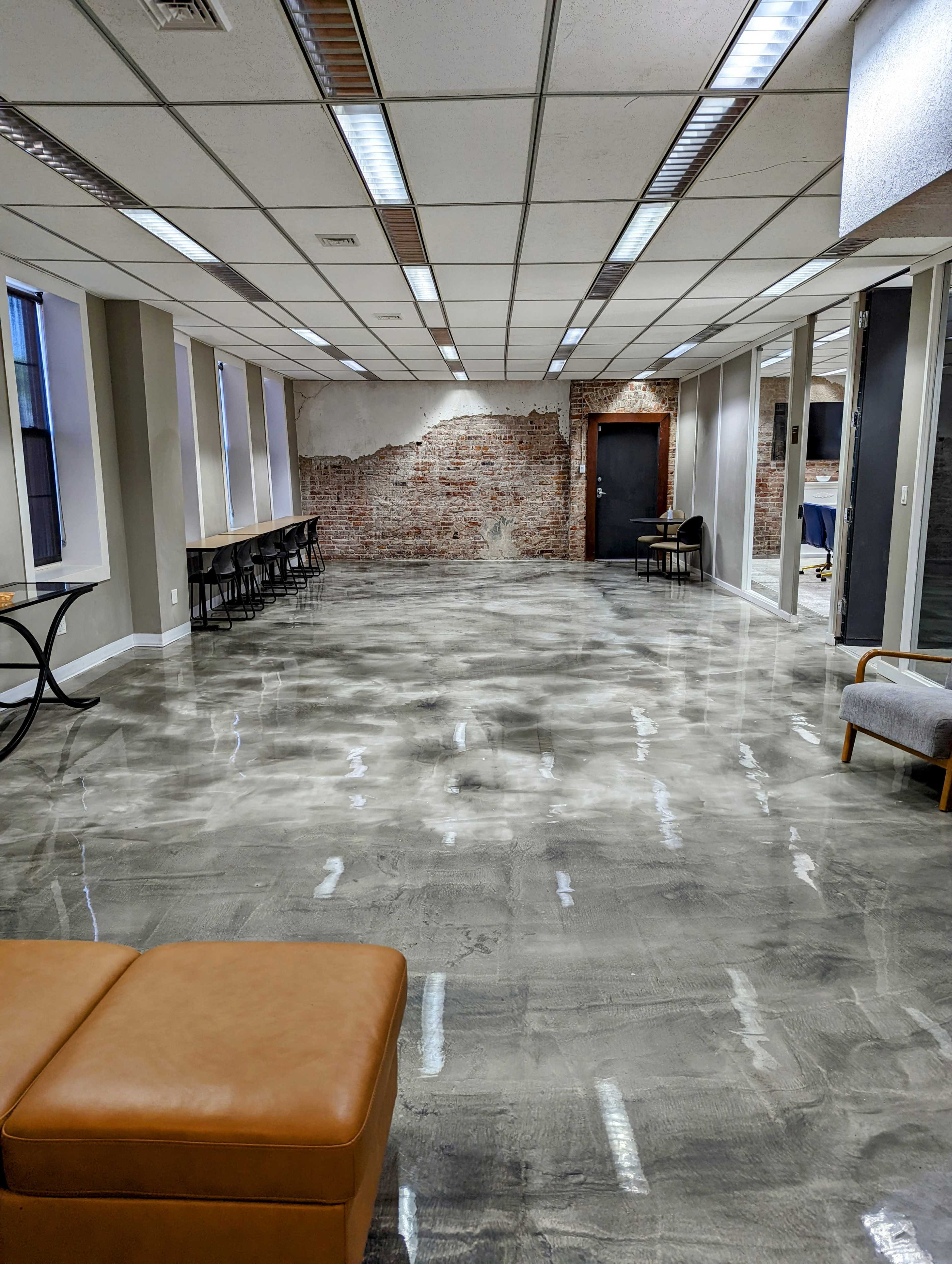 The image shows a spacious interior room with polished concrete flooring, exposed brick walls, and a mix of modern and minimalist furnishings.