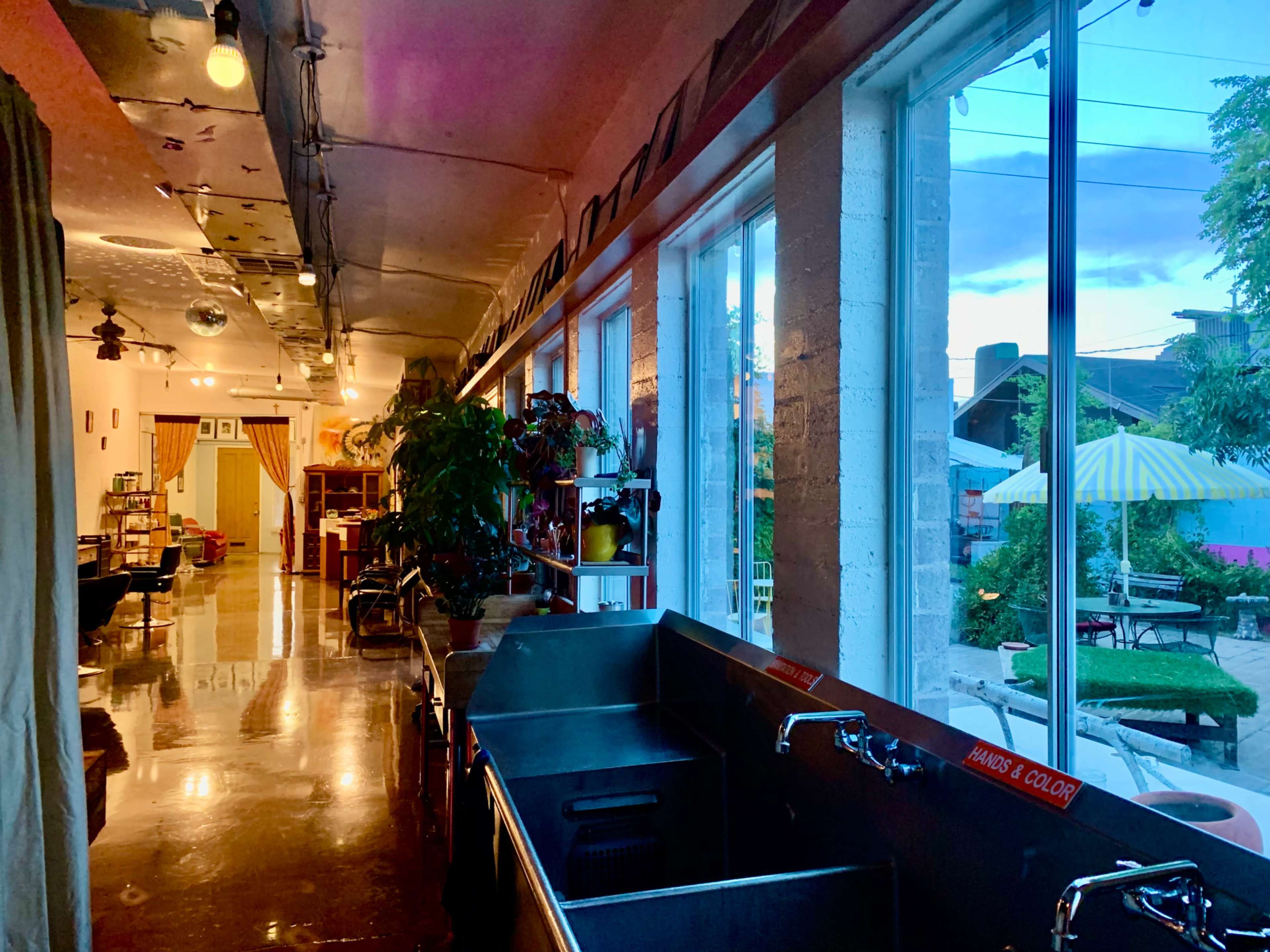 A spacious, well-lit interior features large windows overlooking a garden, with a sink area visible in the foreground.