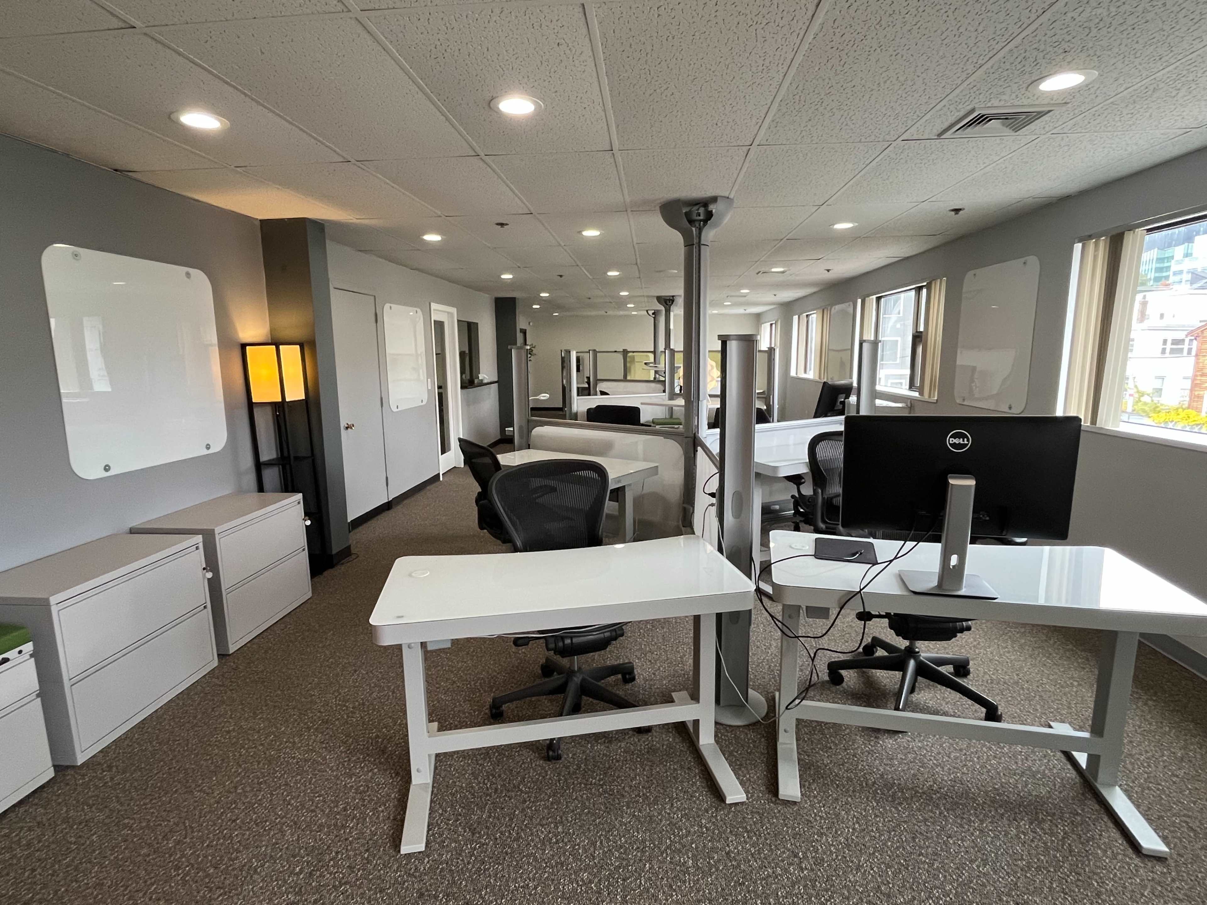 The image shows a modern office space with multiple desks, ergonomic chairs, and large windows, featuring a clean and organized layout.