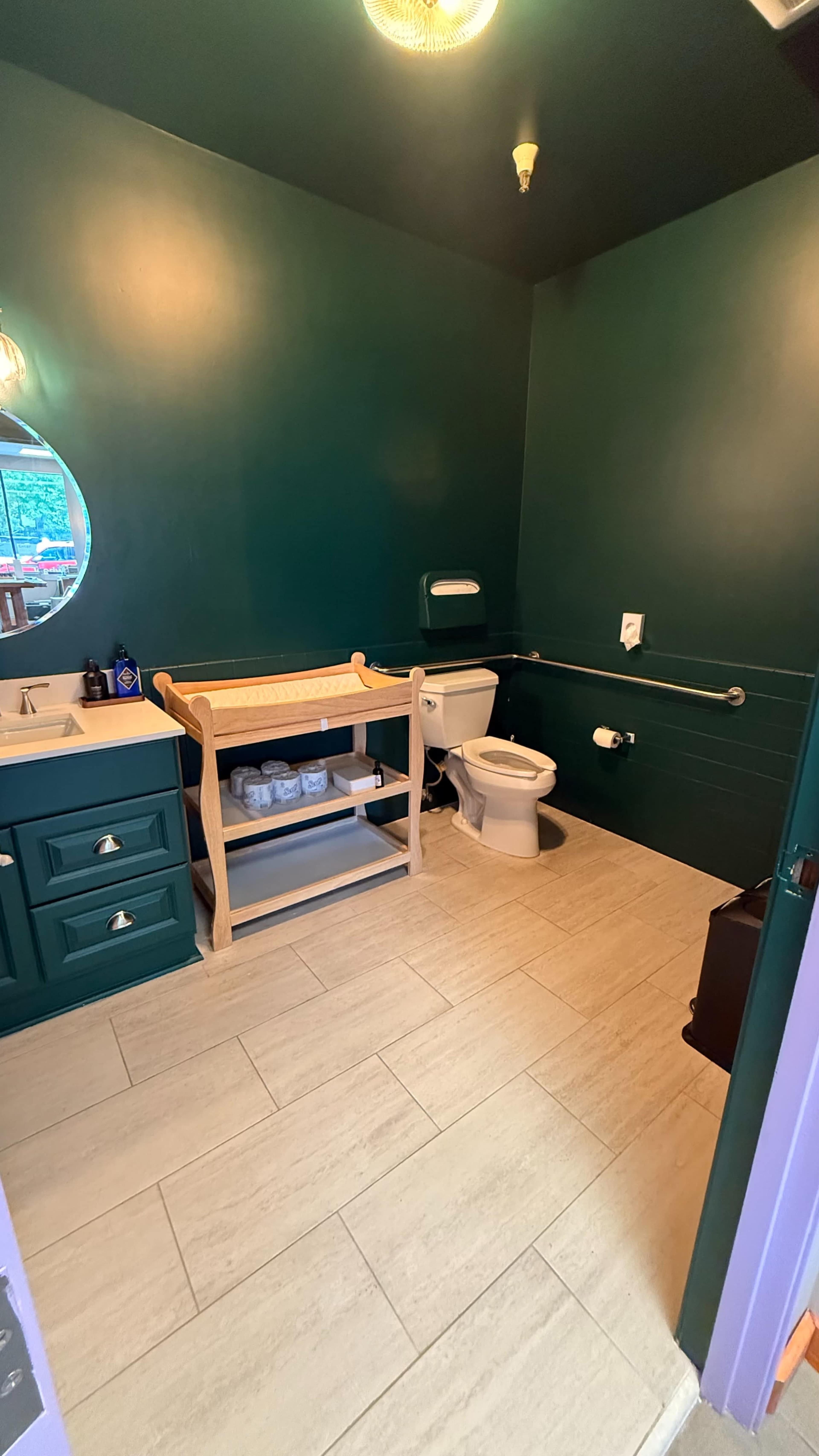 The image shows a small bathroom with dark green walls, a changing table, a toilet, and a sink with a mirror.