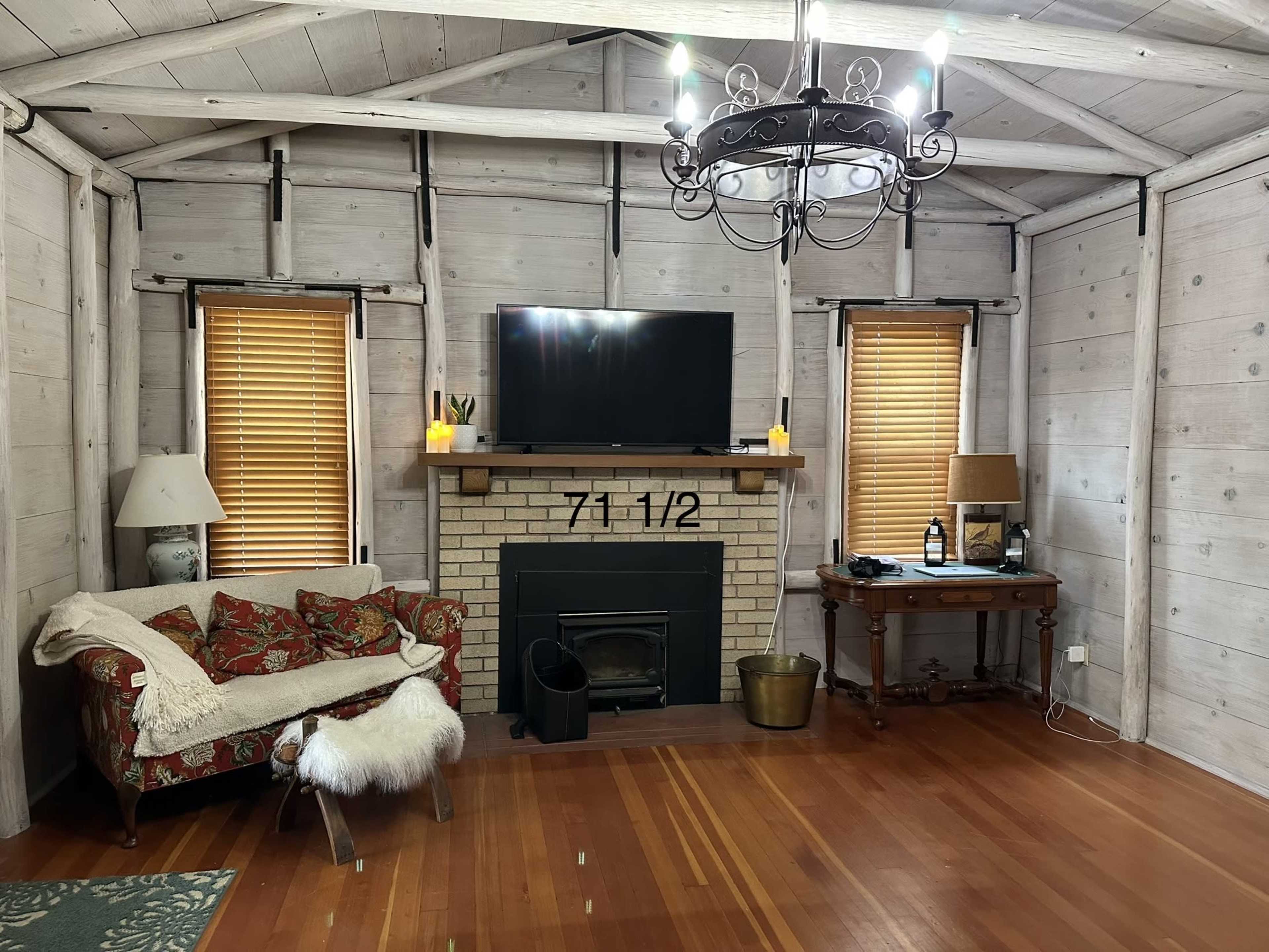 The cozy living room features a brick fireplace with a television mounted above it, flanked by two windows with wooden blinds, and includes a vintage sofa and a small wooden table.