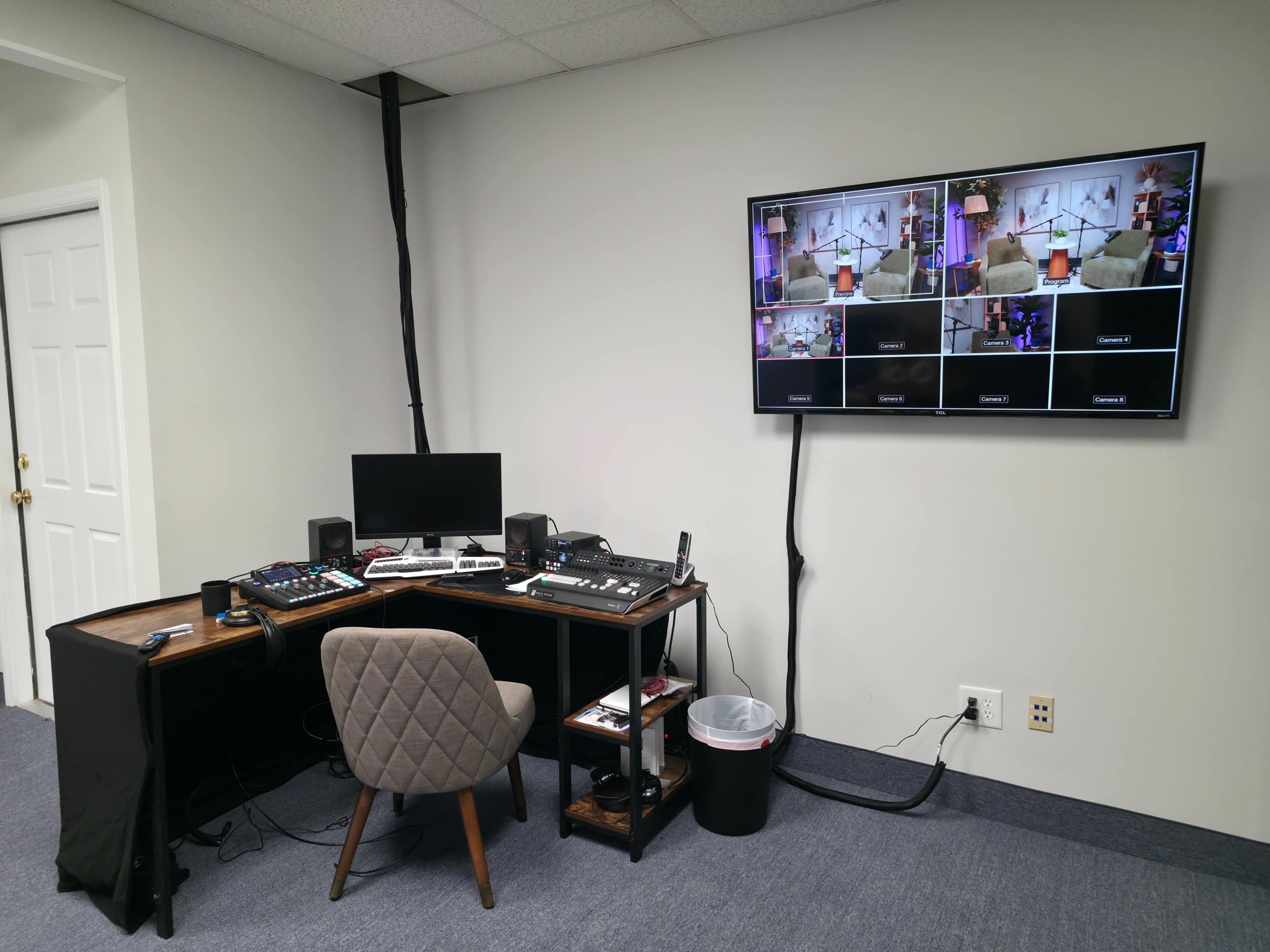 The image shows a small control room setup with a desk, a computer monitor, audio equipment, and a large television displaying multiple video feeds.