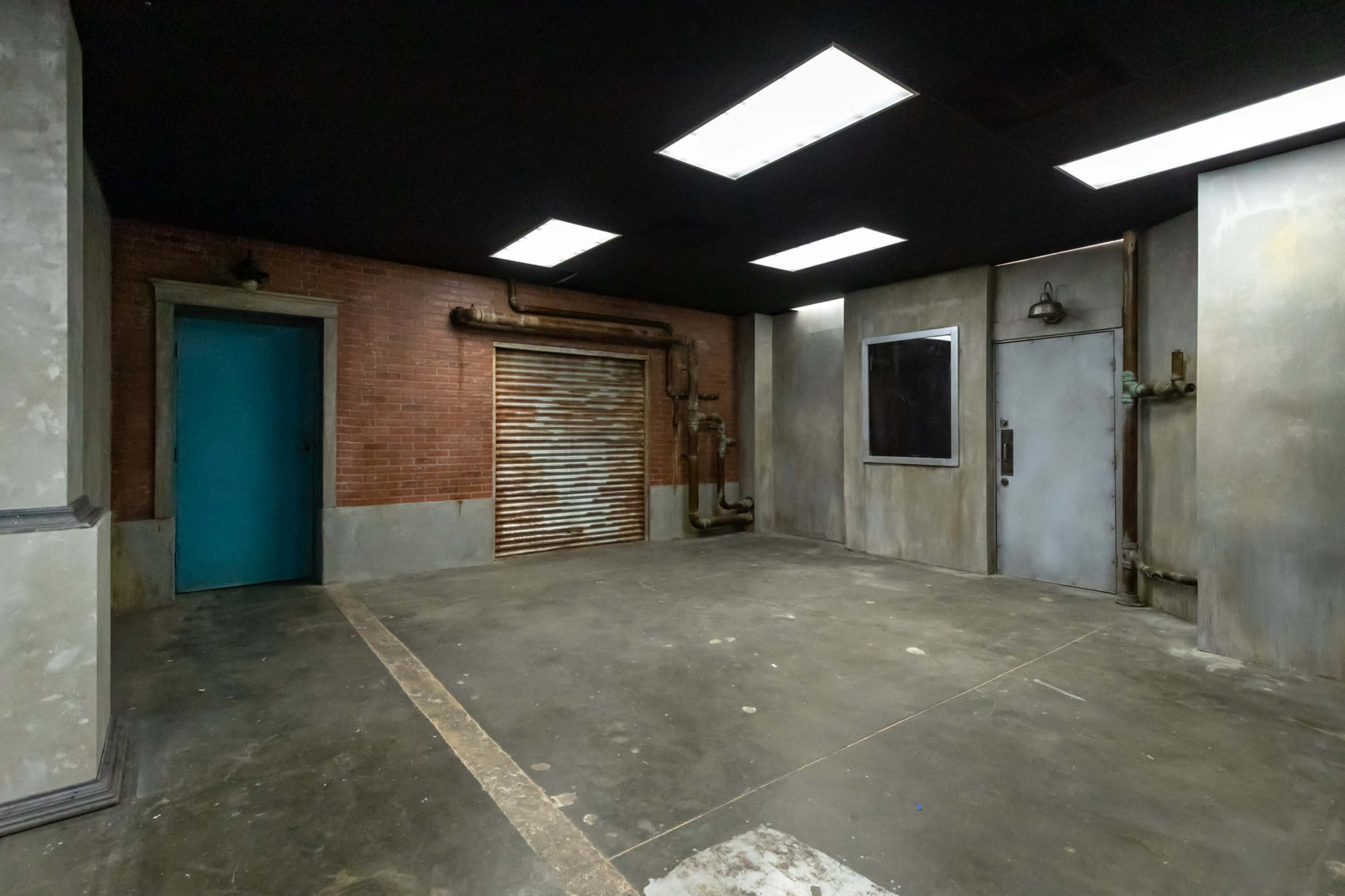 The image shows an empty industrial-style room featuring exposed brick walls, a blue door, and metallic pipes along the surfaces.