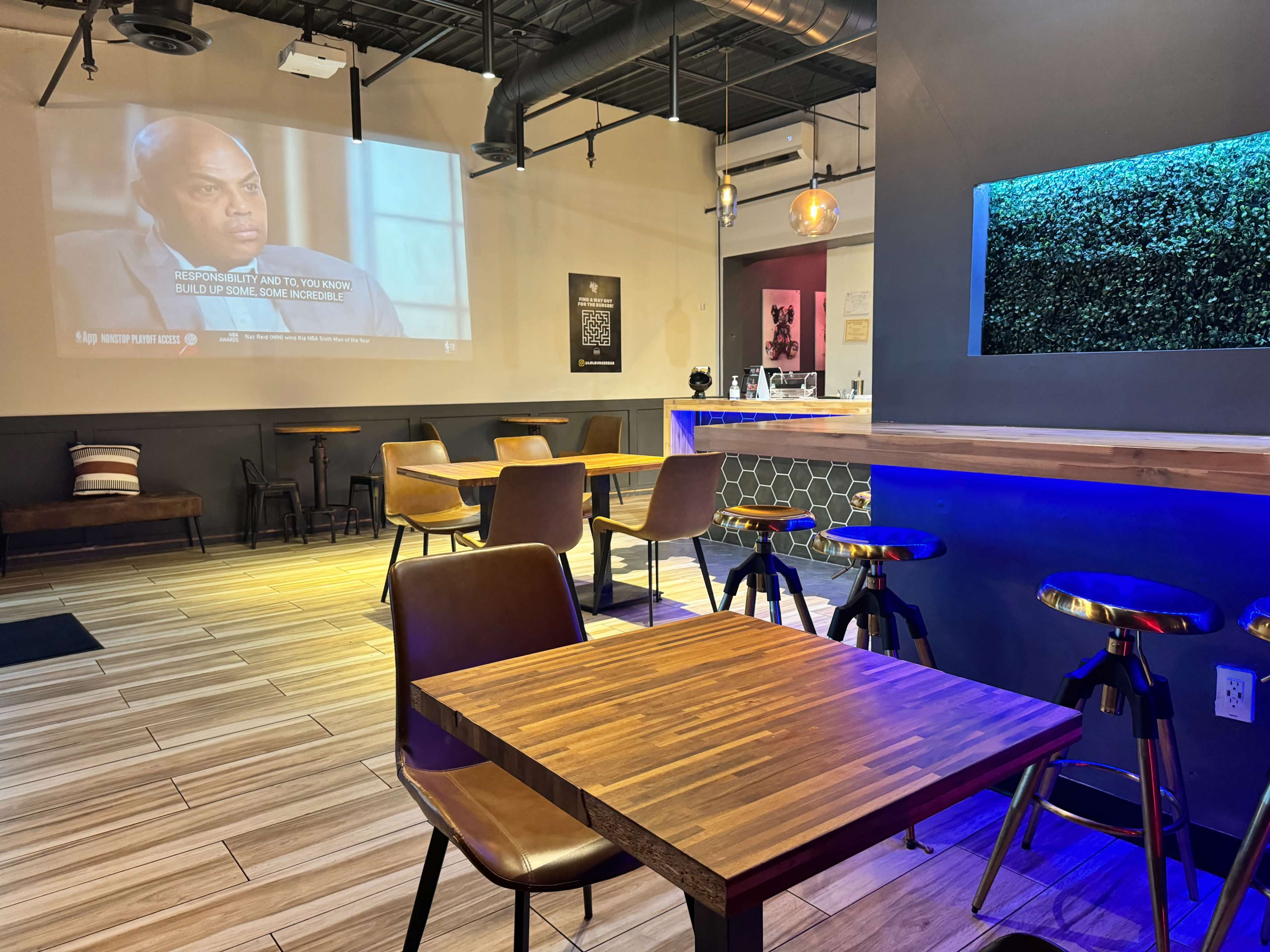 An empty café interior with wooden tables and chairs, a wall-mounted screen displaying a program, and a blue-lit counter area.