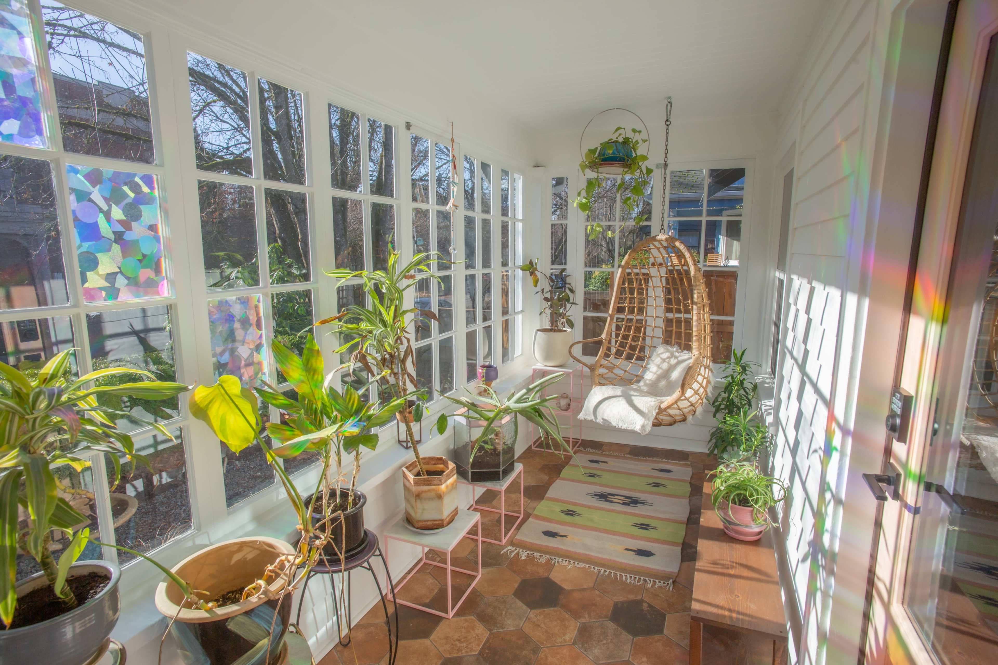 A sunlit sunroom features potted plants, a hanging chair, and large windows that frame the outdoor view.