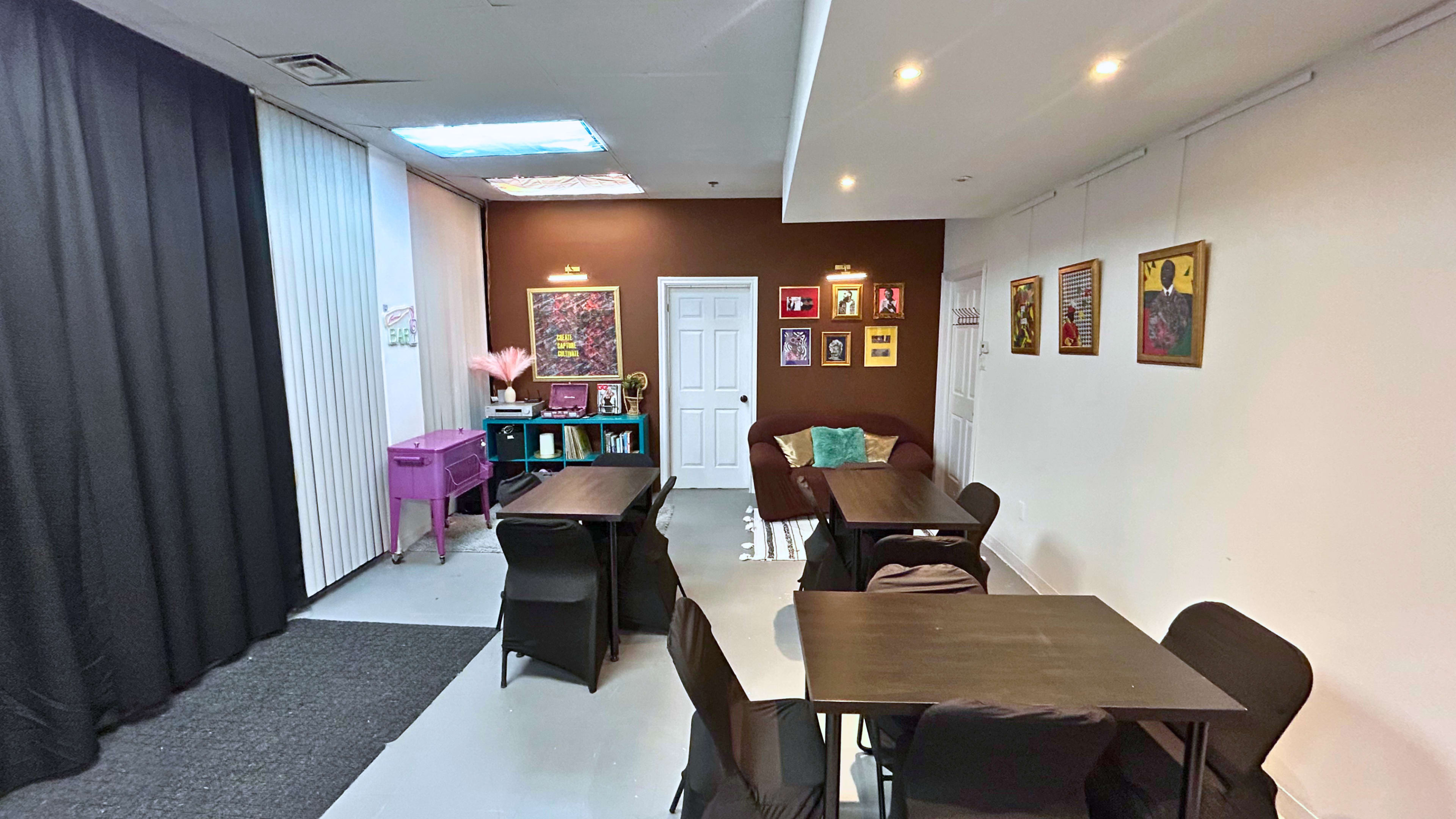 The image shows a spacious room with tables and chairs set for a meeting, featuring a couch, artwork on the walls, and a partitioned area with curtains.
