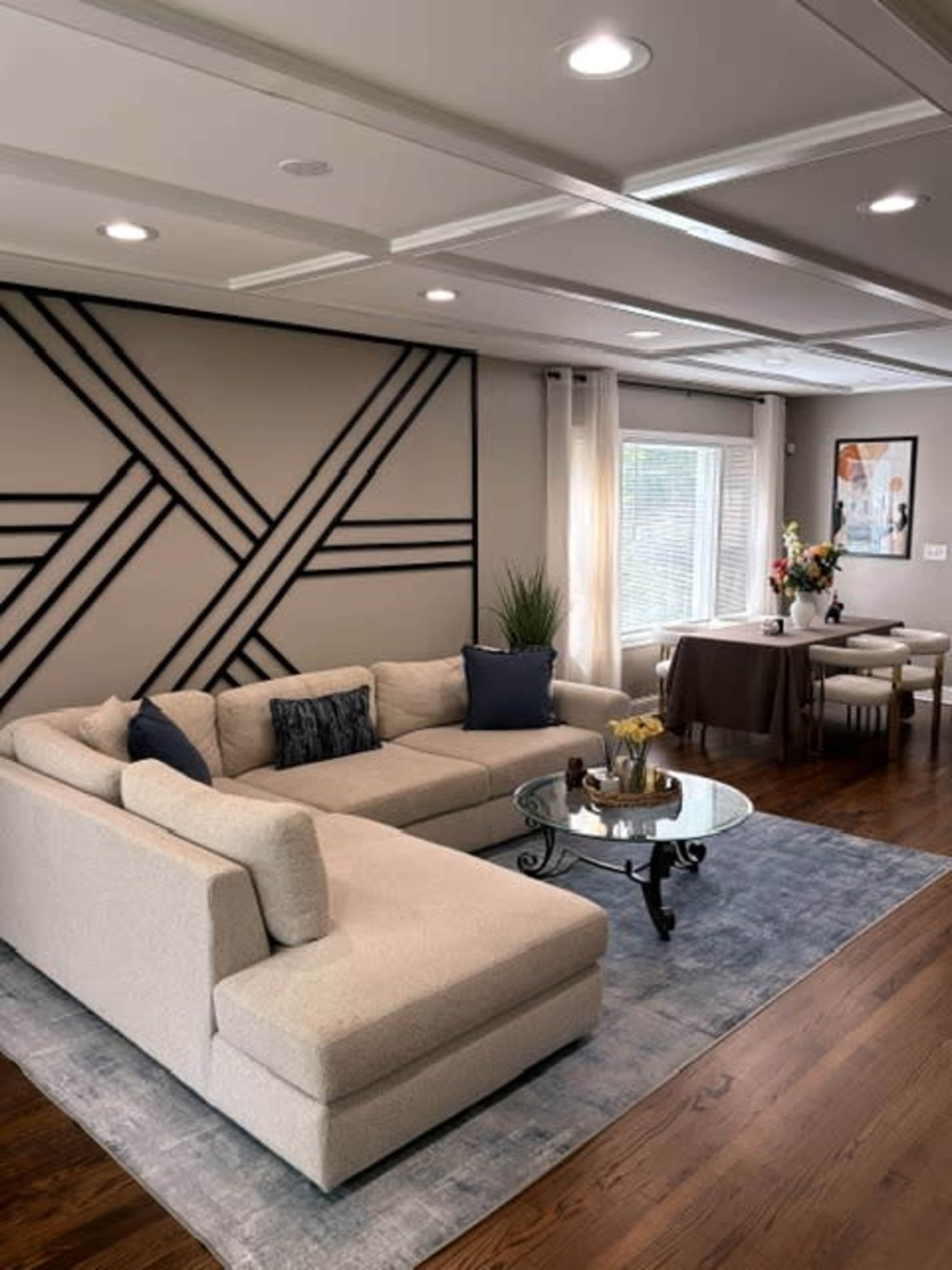 The image shows a modern living room featuring a light-colored sectional sofa, a round glass coffee table, a decorative rug, and a dining area with a small table and chairs.