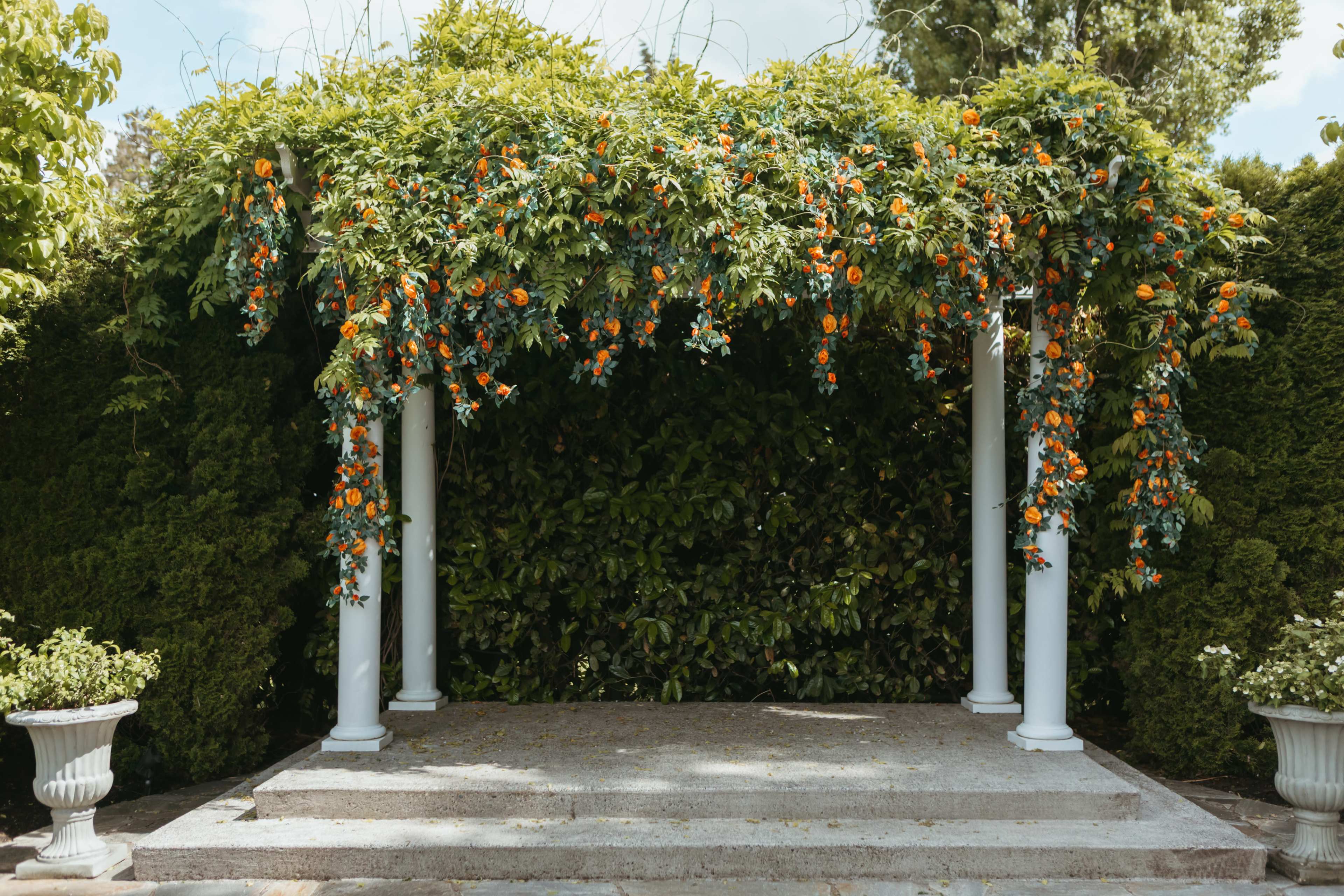 A floral-covered arbor with orange flowers and greenery stands in front of a lush hedge, positioned on a stone platform surrounded by planters.